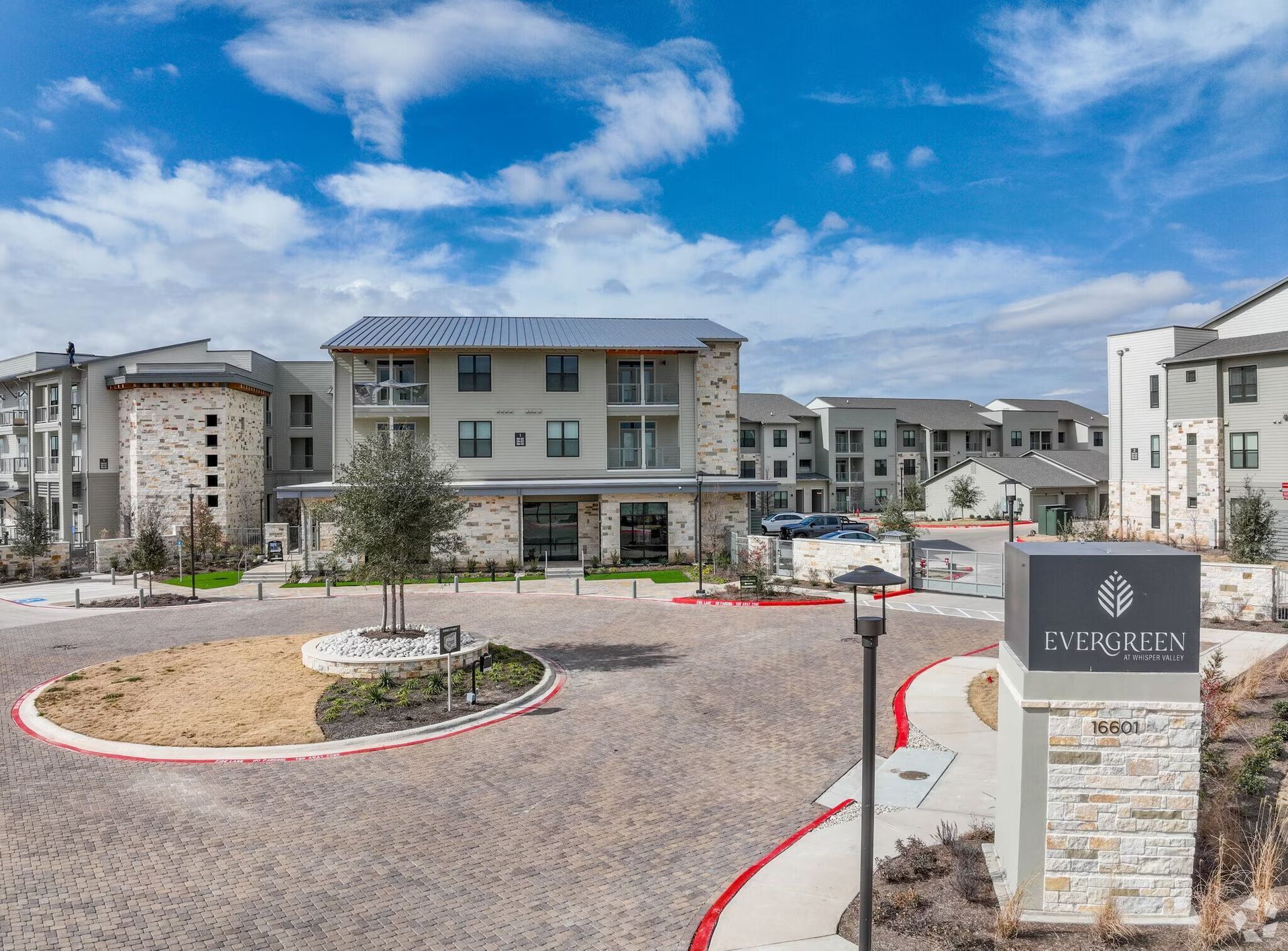 Apartment complex entrance with circular driveway, stone sign, and blue sky