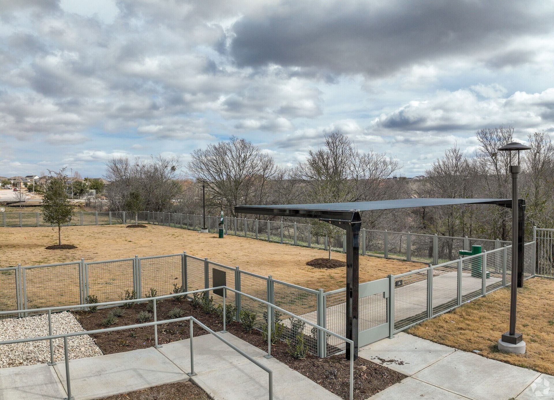 Outdoor park with paved paths, black pergola, fencing, leafless trees, and a cloudy sky.