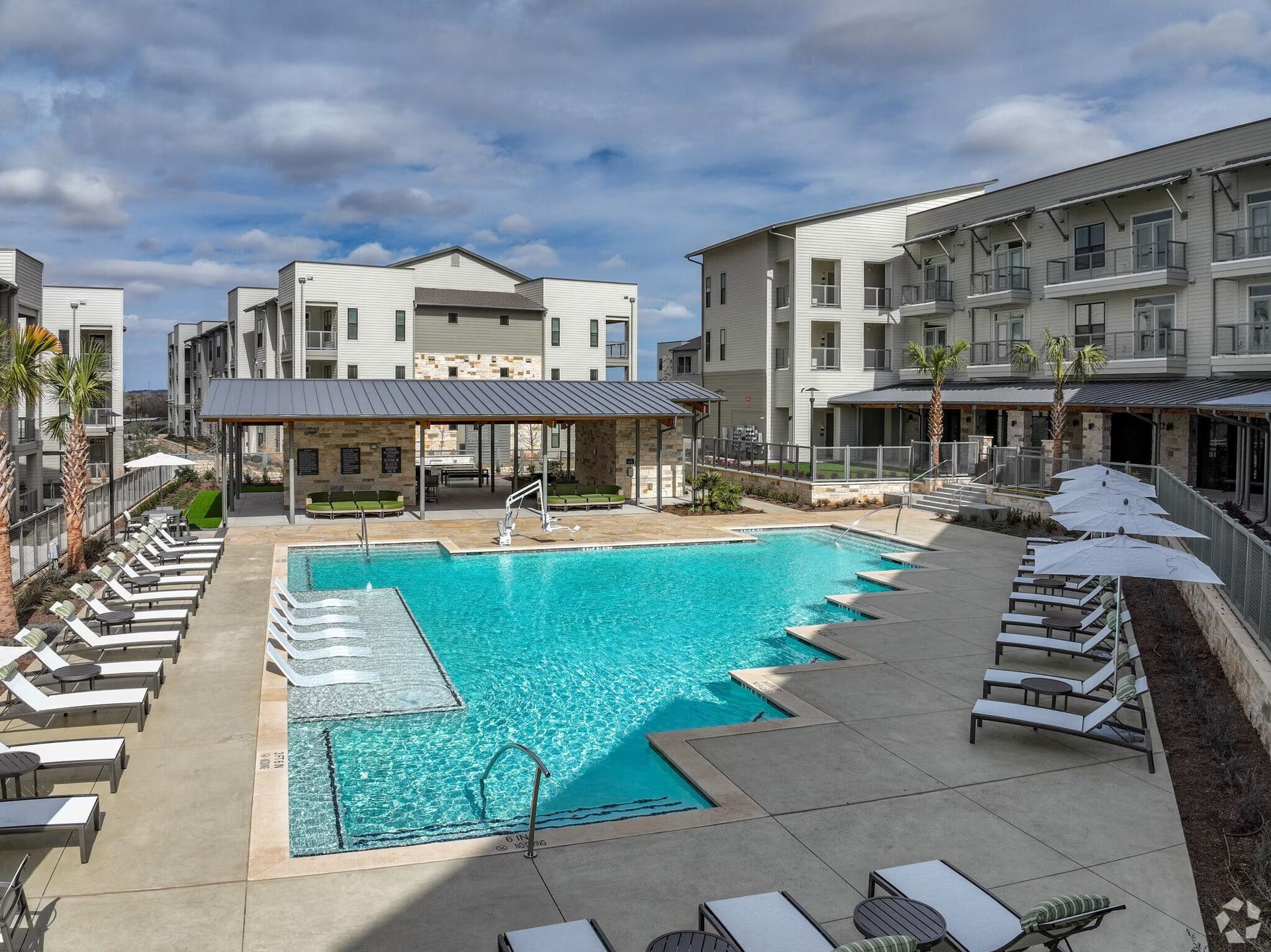 Modern apartment pool courtyard with turquoise water, lounge chairs, and white umbrellas under a cloudy sky