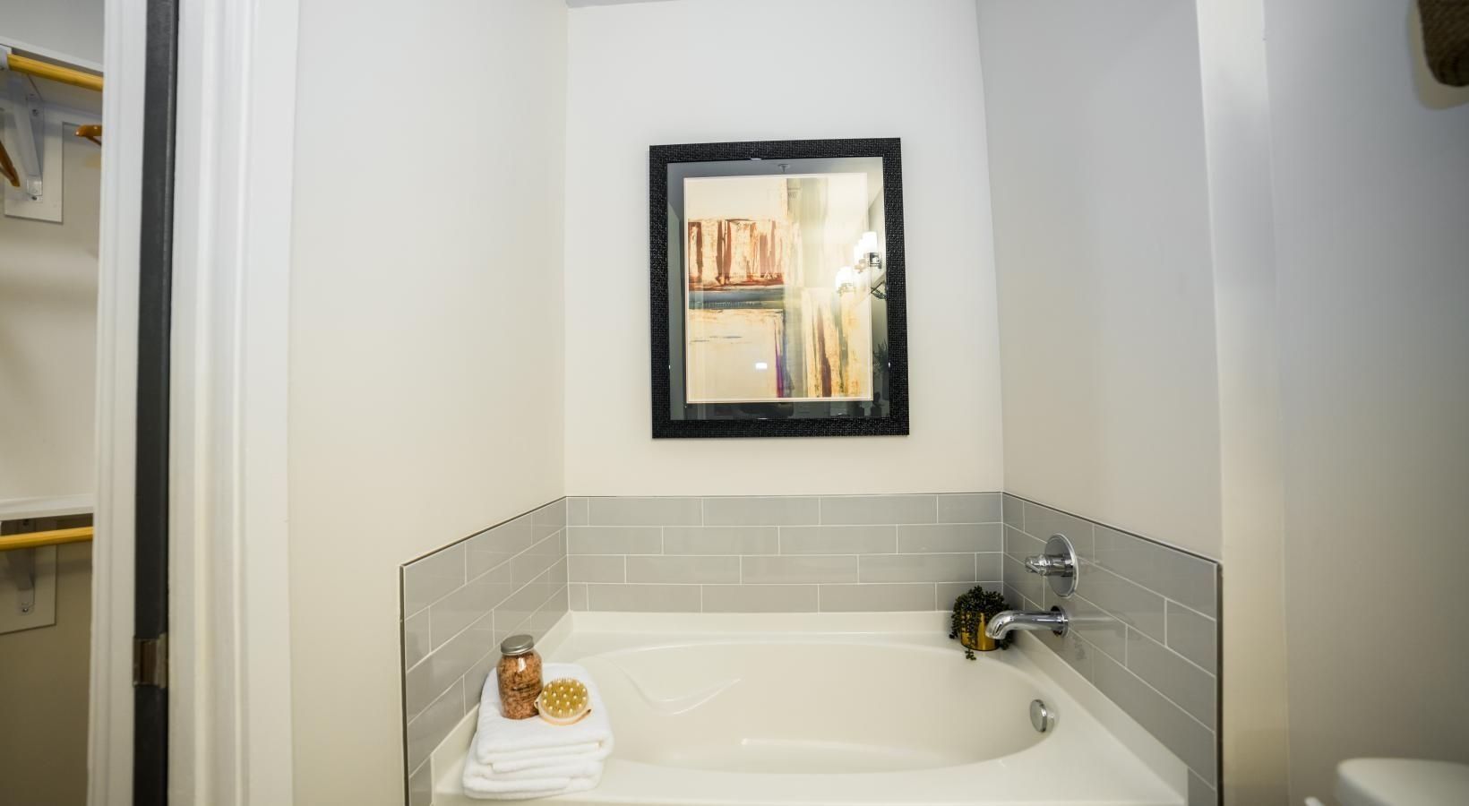 Small white bathroom with a corner bathtub, gray tile, and a framed picture above the tub.