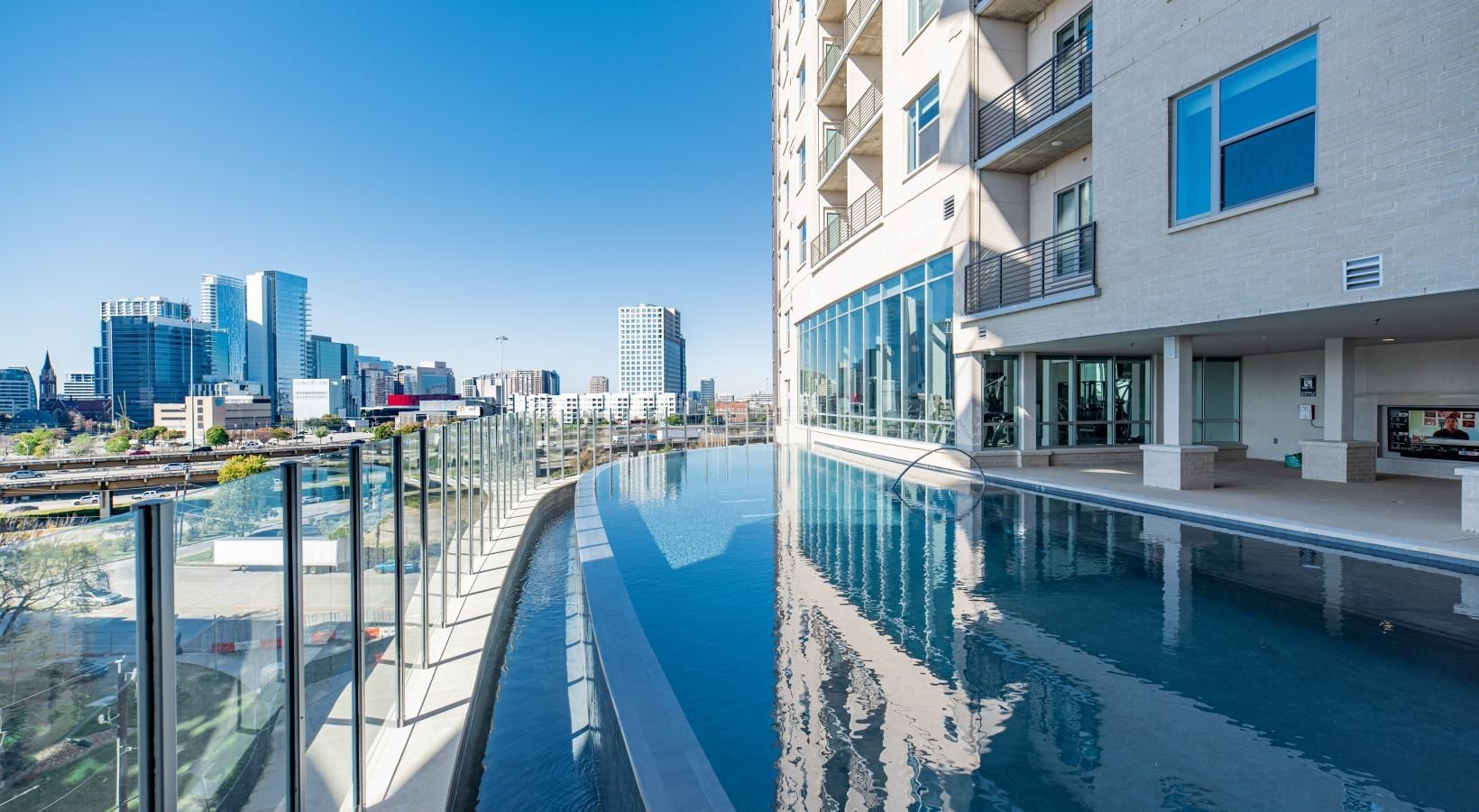 Rooftop infinity pool beside a modern building, reflecting a city skyline under a bright sky