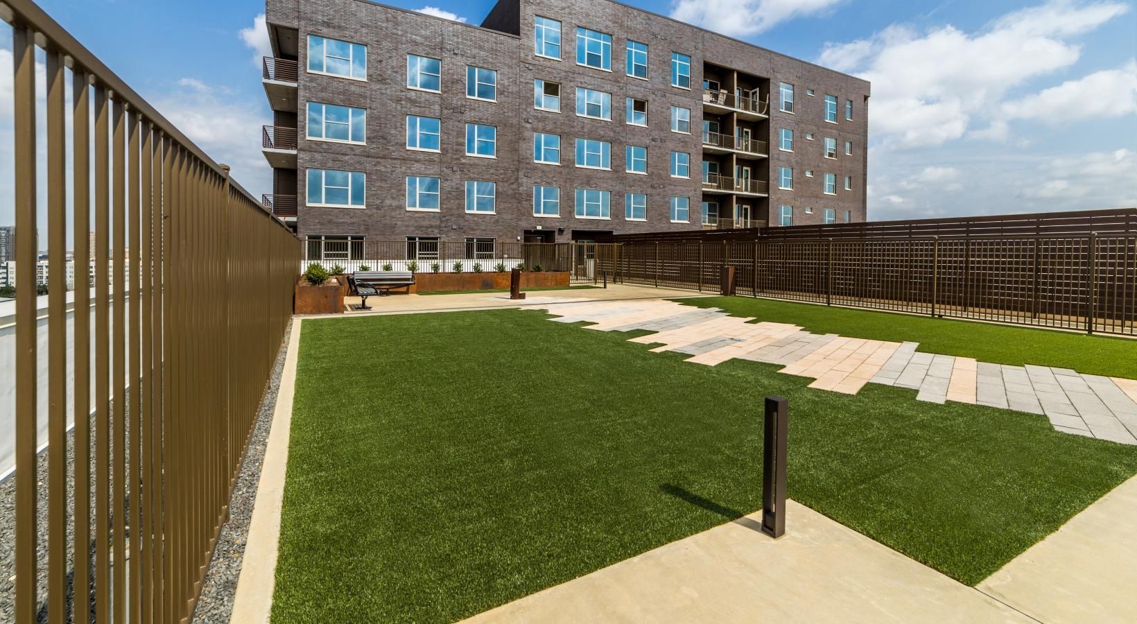 Courtyard with green lawn, paved paths, wooden fence, and a brick apartment building in the background