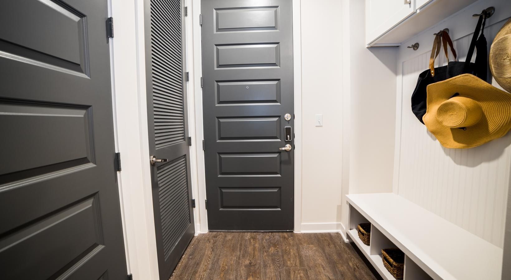 Narrow entryway with gray front door, white walls, bench, coat hooks, and a yellow hat hanging on the right
