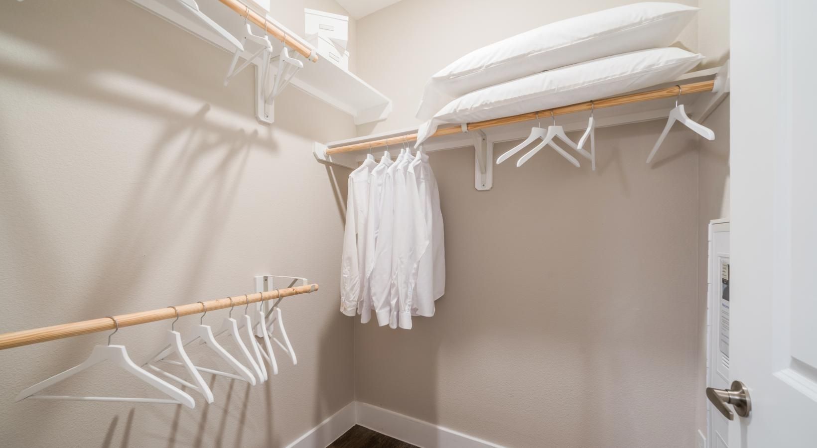 Empty walk-in closet with beige walls, white shelves, hanging rods, and a few clothes hangers and pillows.