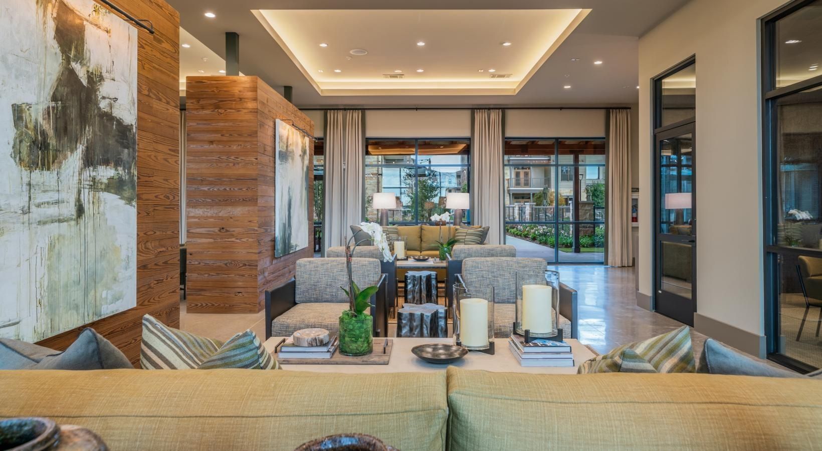 Modern living room with beige sofas, wood paneling, large artwork, and windows opening to a patio.