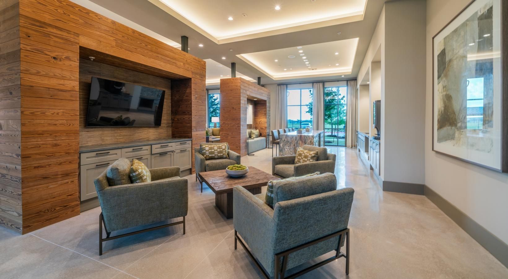 Modern apartment lobby with lounge chairs, wall-mounted TV, fireplace, and large windows overlooking greenery