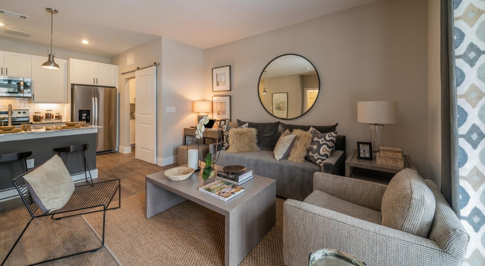Modern living room and kitchen with gray furniture, round mirror, rug, and pendant lights