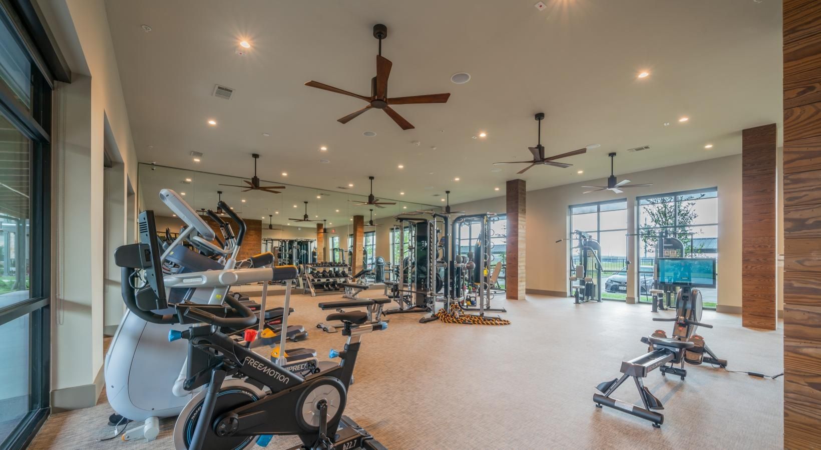 Modern gym interior with cardio and weight equipment, ceiling fans, and large windows