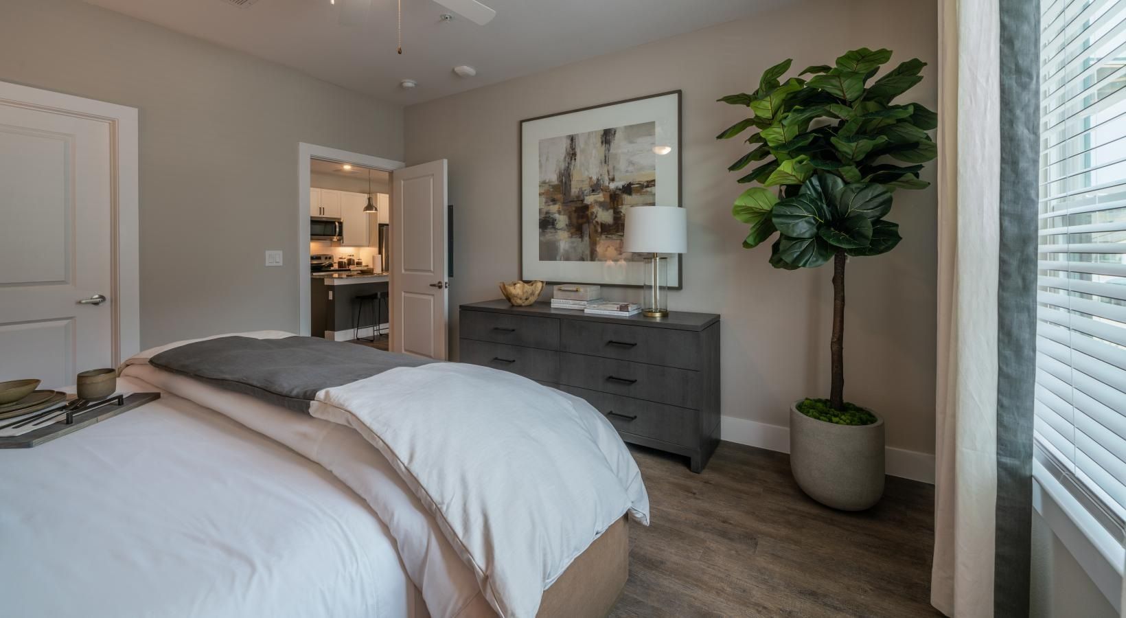 Bright bedroom with a bed, gray dresser, lamp, framed art, and a potted plant beside the window.