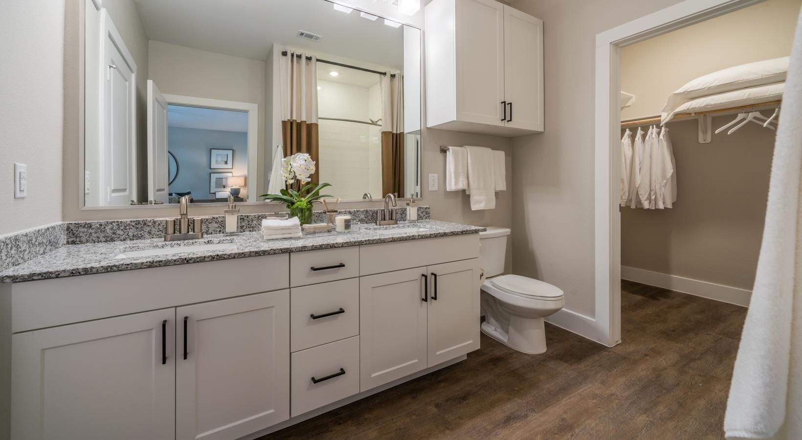 Bright bathroom vanity with double sinks, large mirror, toilet, and open walk-in closet nearby.