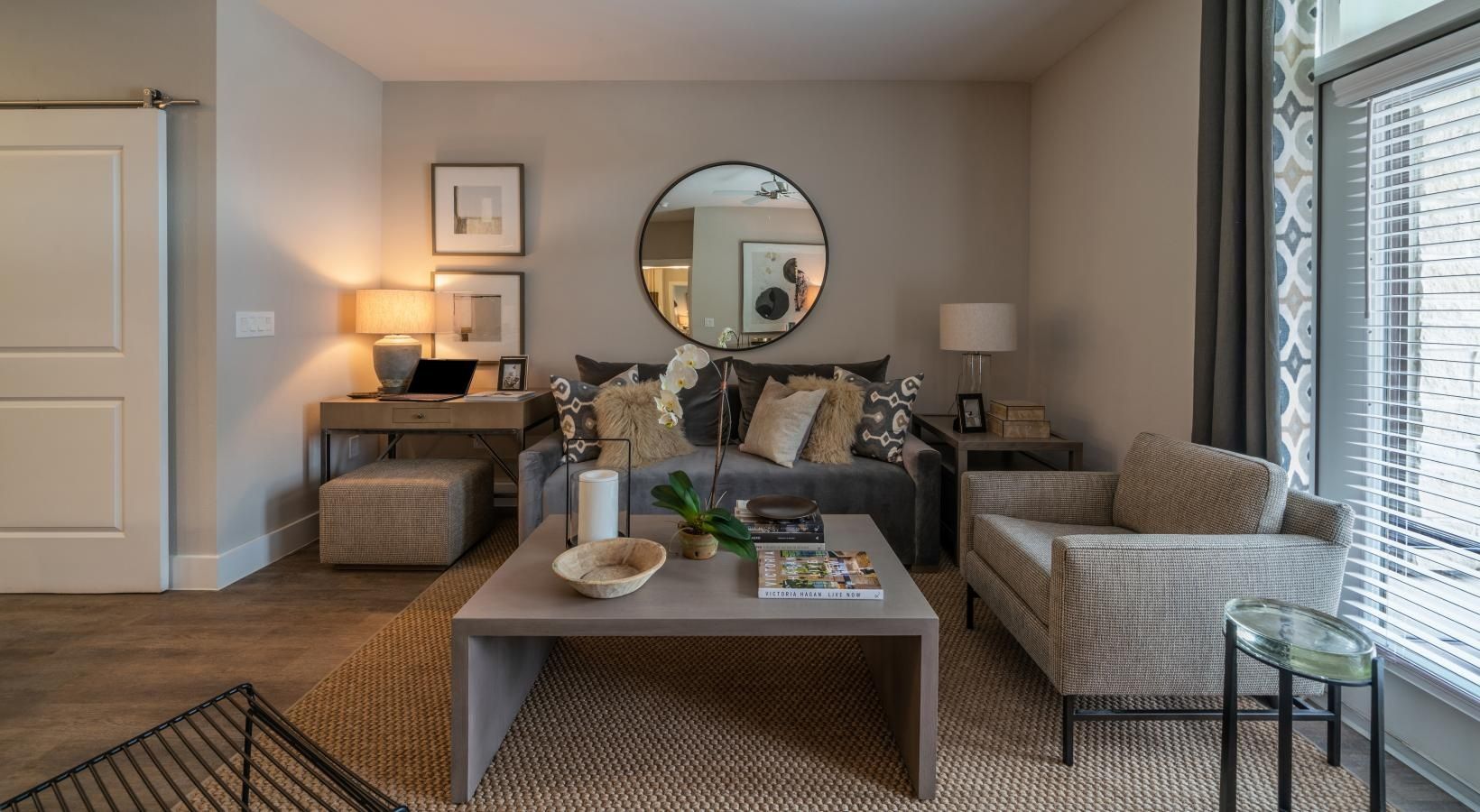 Modern living room with beige sofa, coffee table, round mirror, floor lamp, and window blinds
