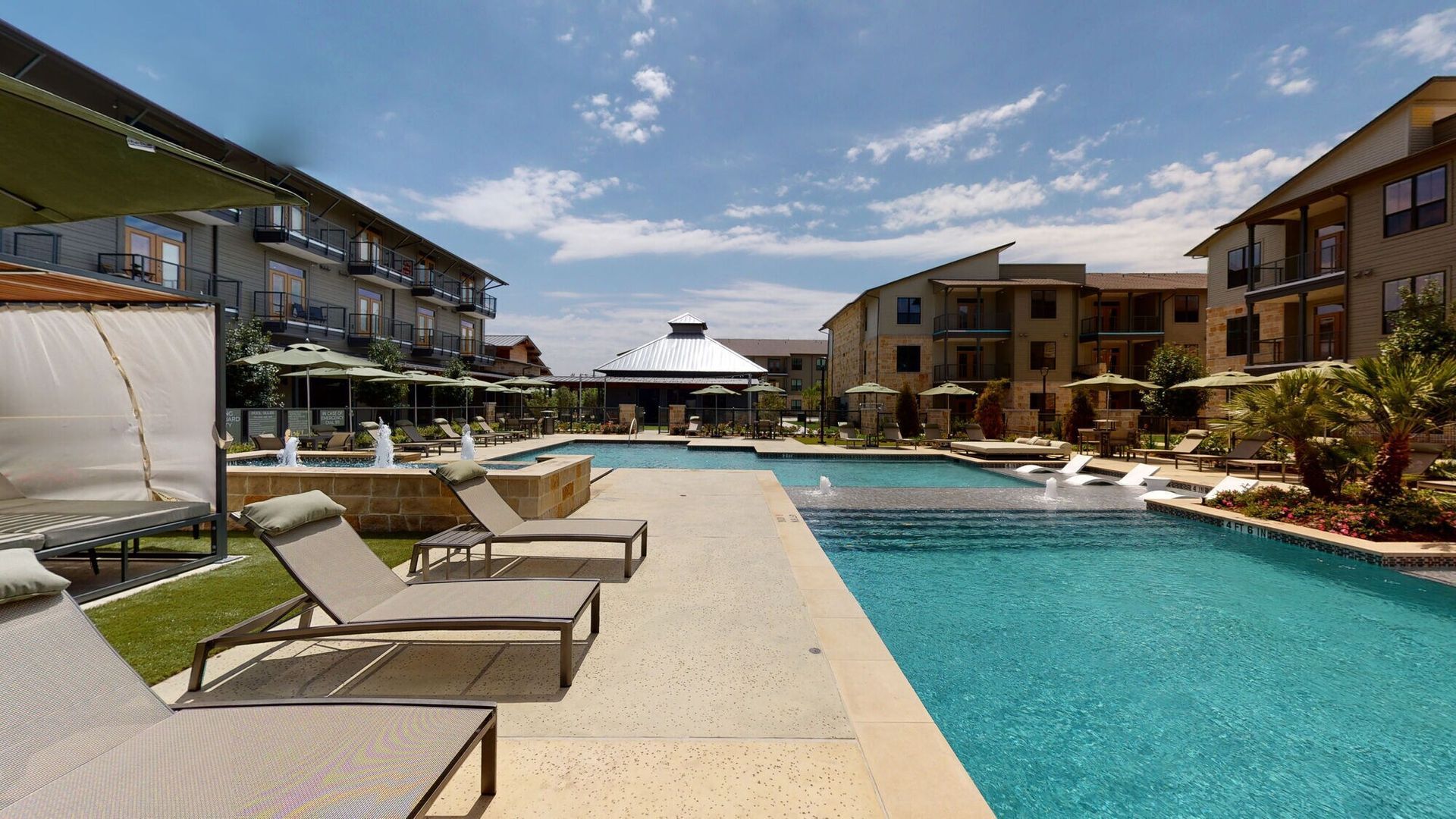 Resort pool with lounge chairs beside turquoise water and surrounding hotel buildings under a blue sky