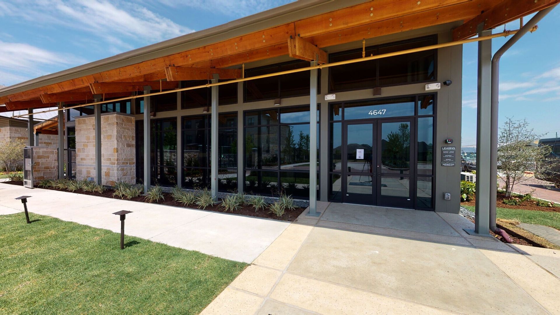 Modern building entrance with glass doors, wood overhang, and a concrete walkway beside a lawn.