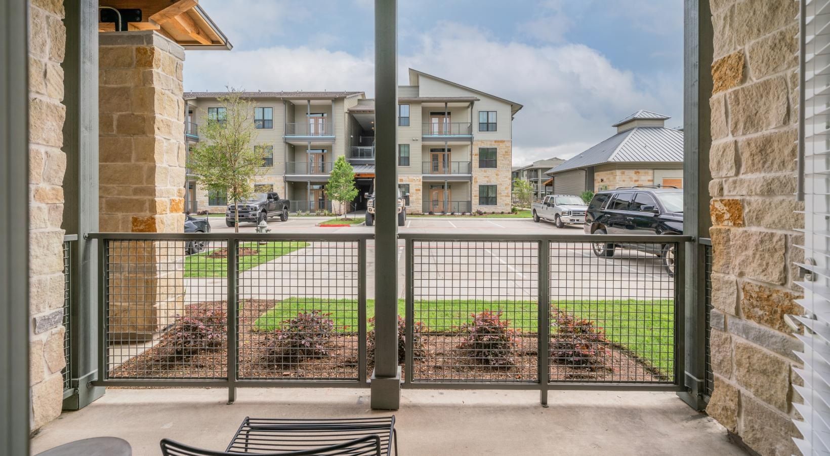 Covered apartment patio with stone columns, black railing, and view of neighboring buildings across a street.