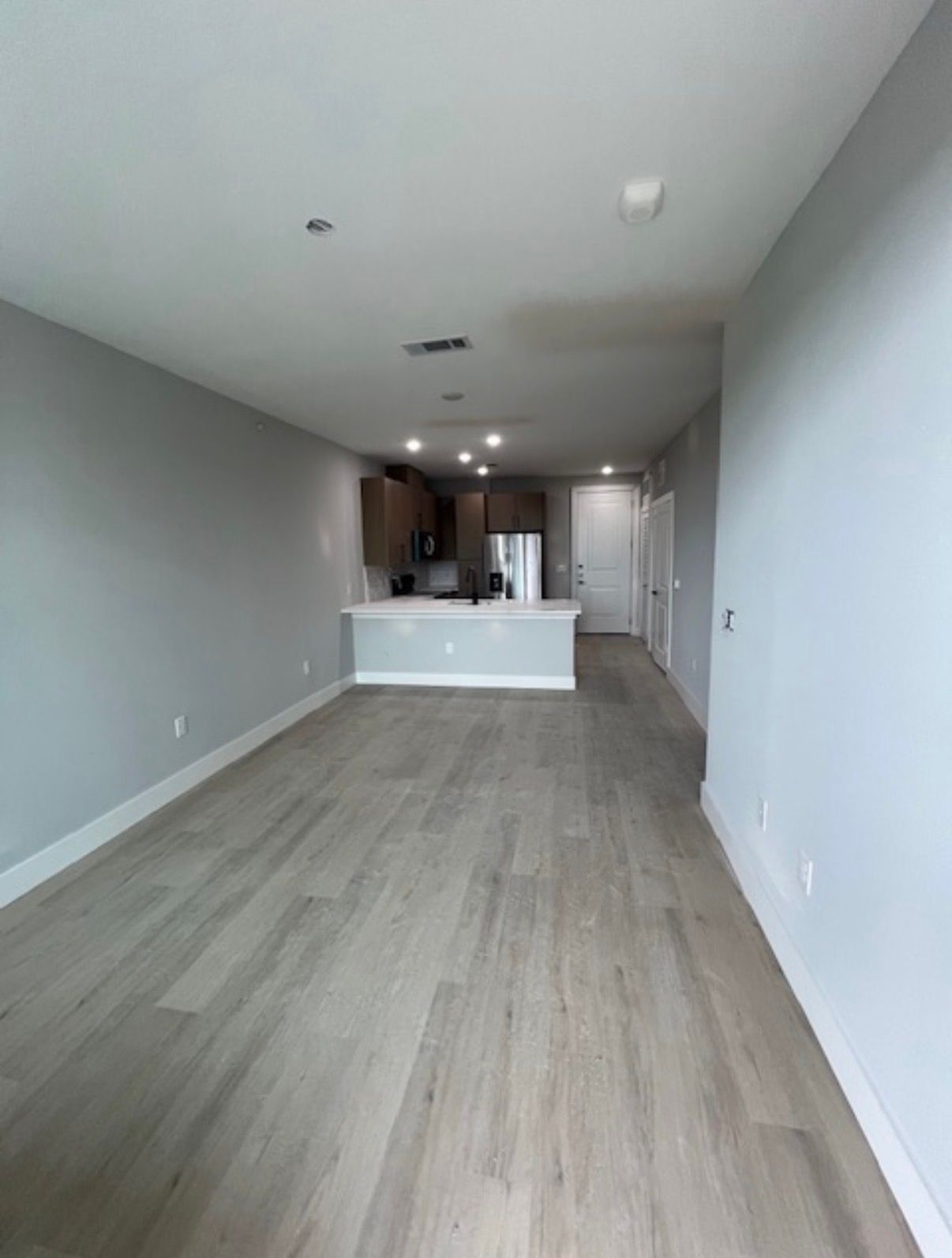 Empty modern apartment hallway with light wood floors, white walls, and a kitchen at the far end