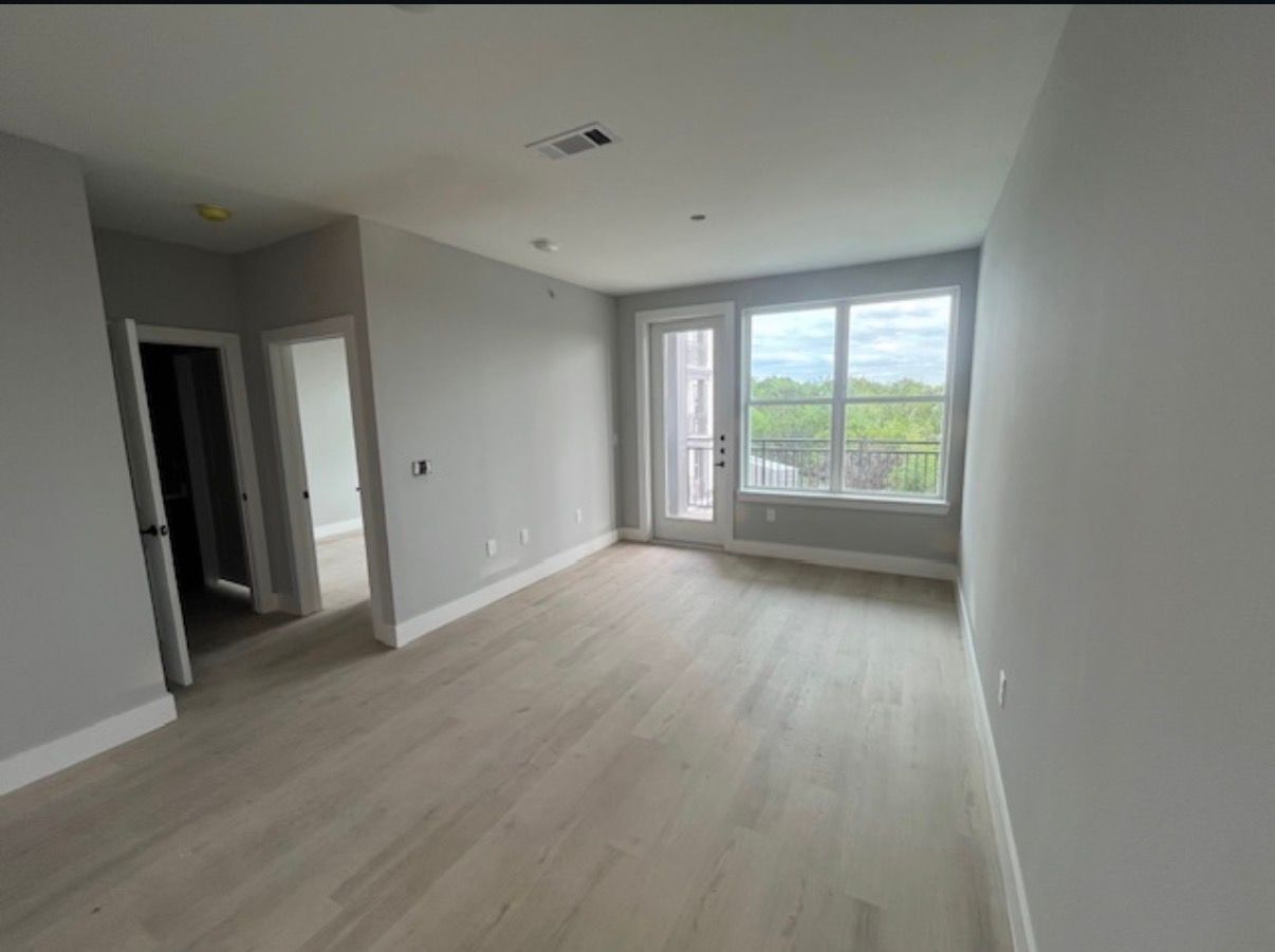 Empty unfurnished room with light wood floors, gray walls, and a large window overlooking greenery