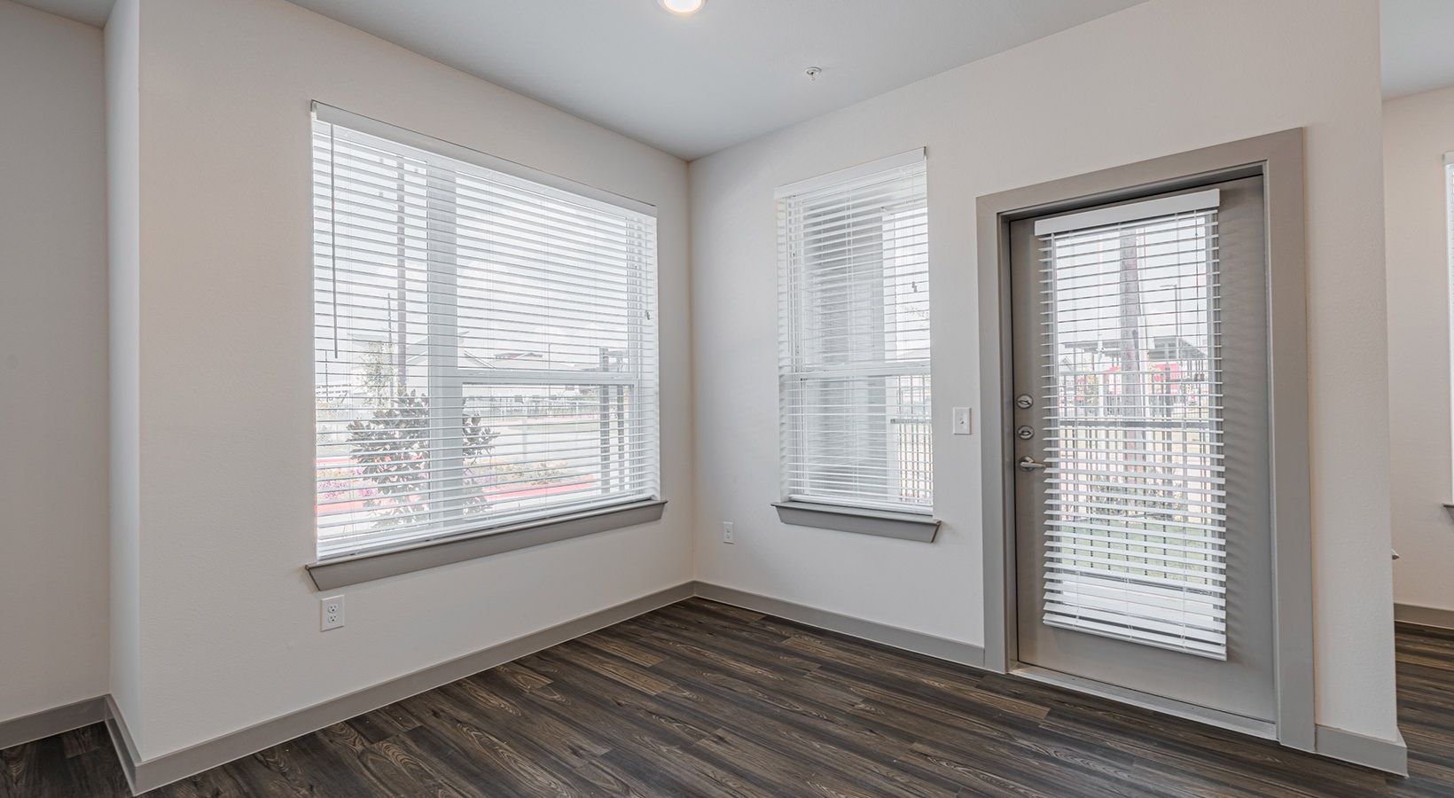 Empty modern room with white walls, gray wood floors, and windows with blinds and a glass door