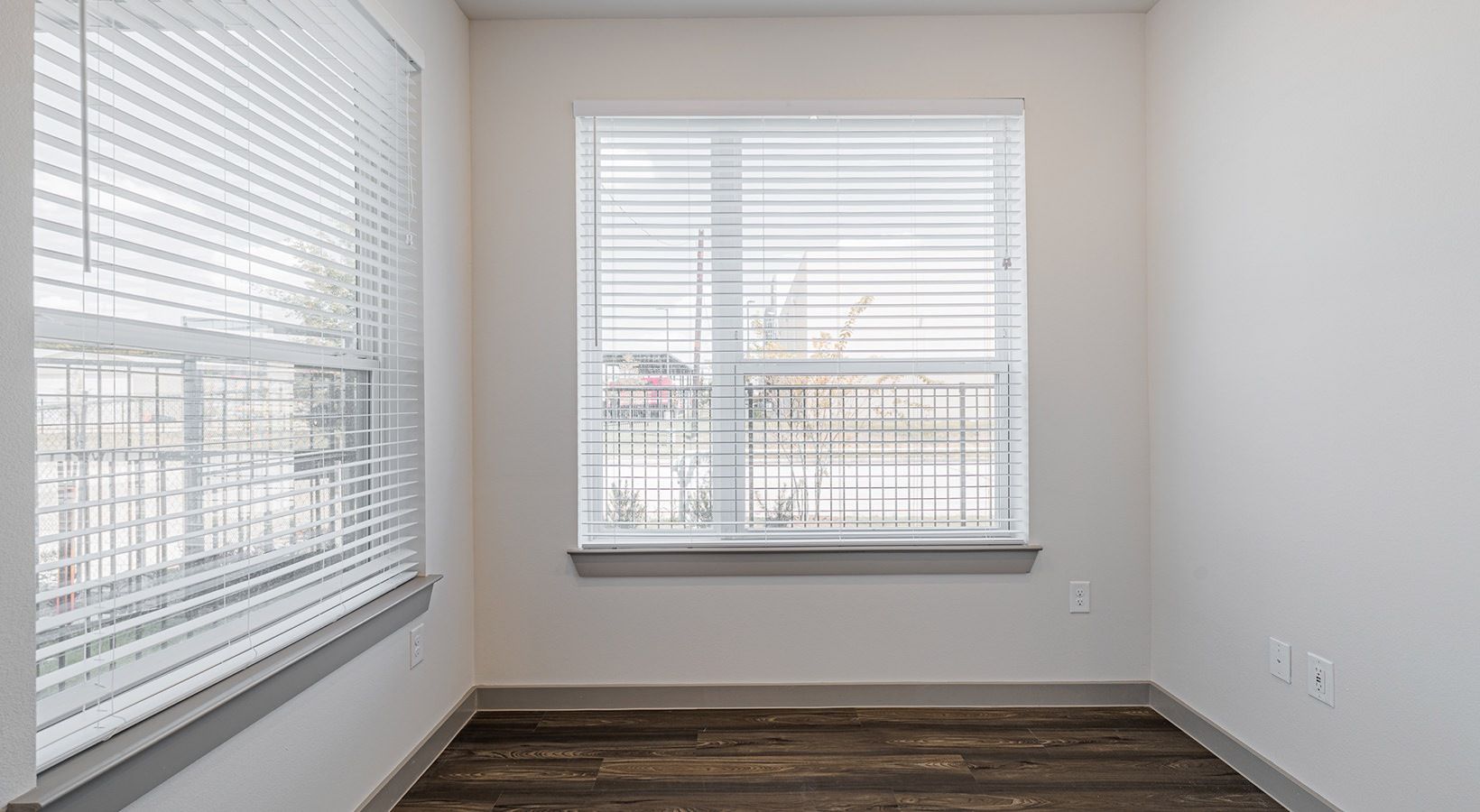 Empty room with two windows, white blinds, and dark wood flooring