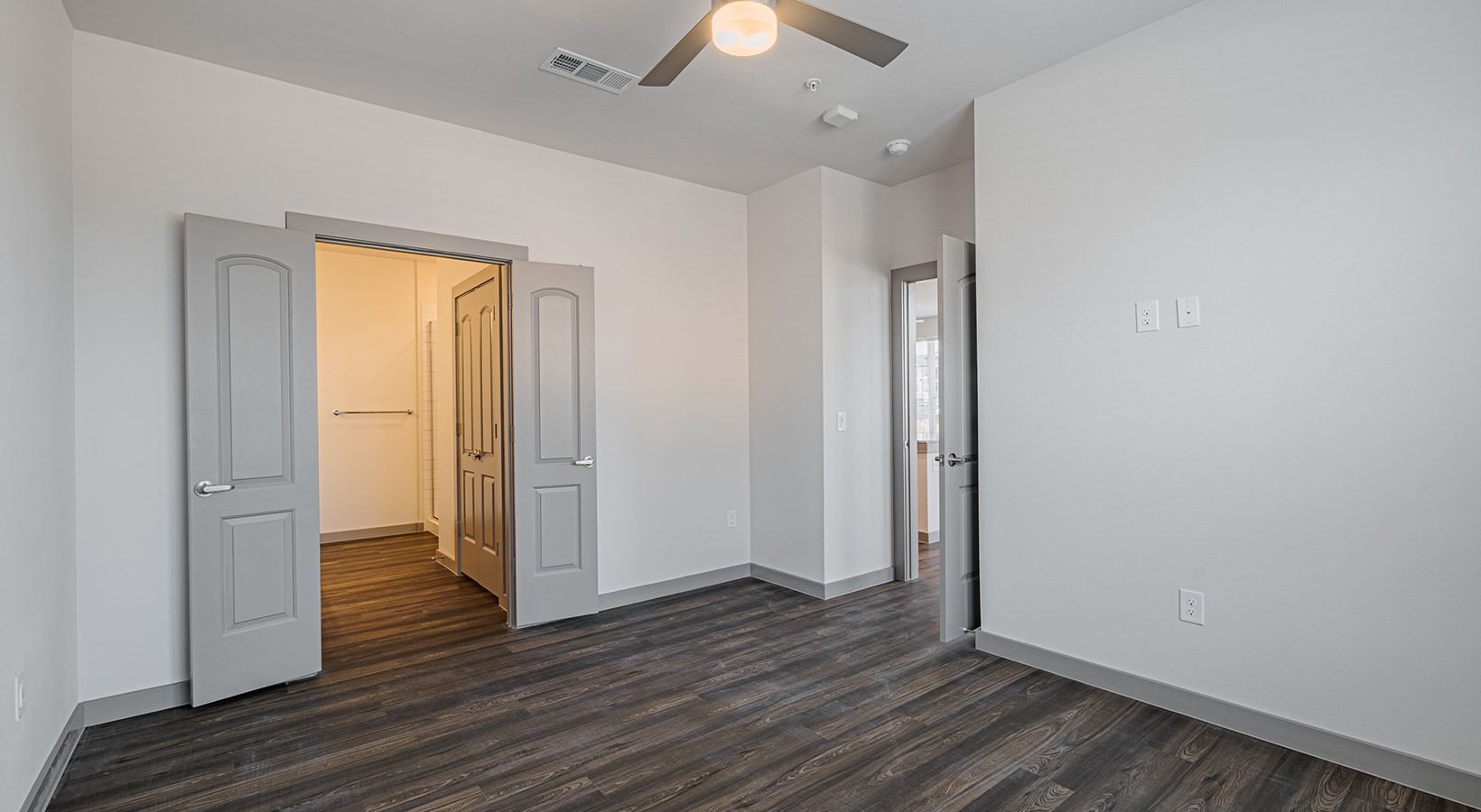 Empty bedroom with gray walls, wood-look flooring, white doors, and a ceiling fan.