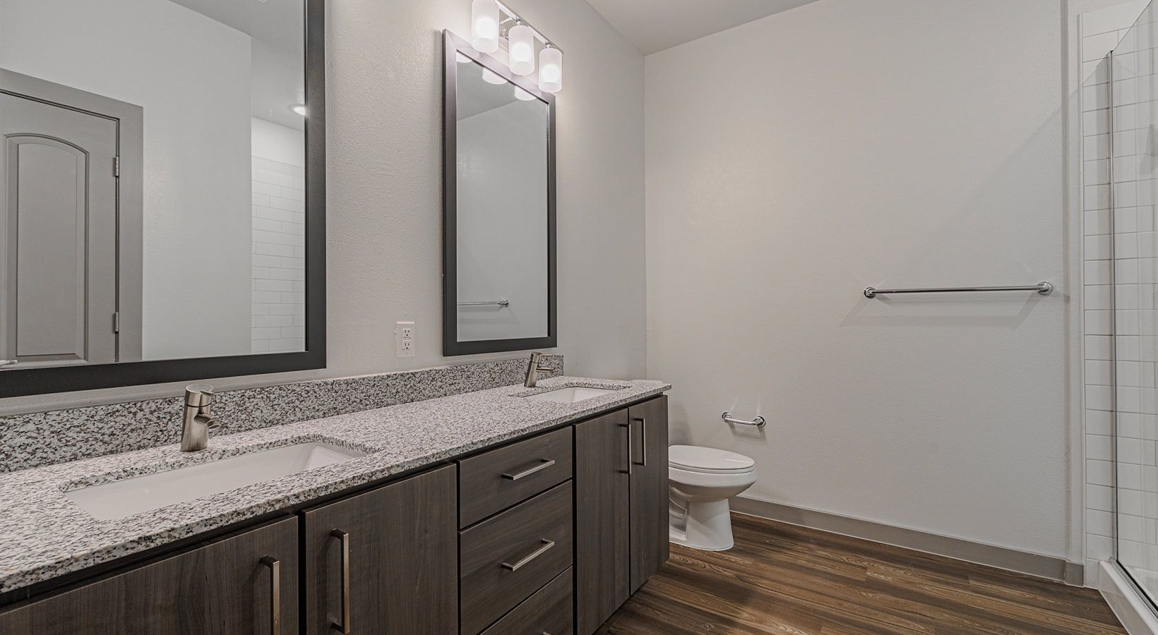 Modern bathroom with double vanity, mirrors, gray cabinets, white walls, toilet, and wood-look floor.