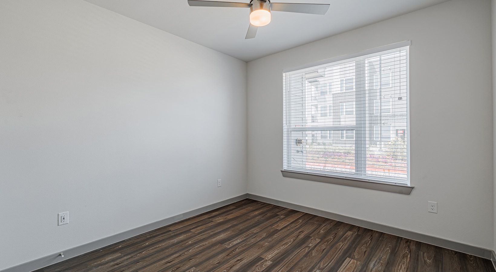 Empty bedroom with white walls, wood flooring, a window with blinds, and a ceiling fan.