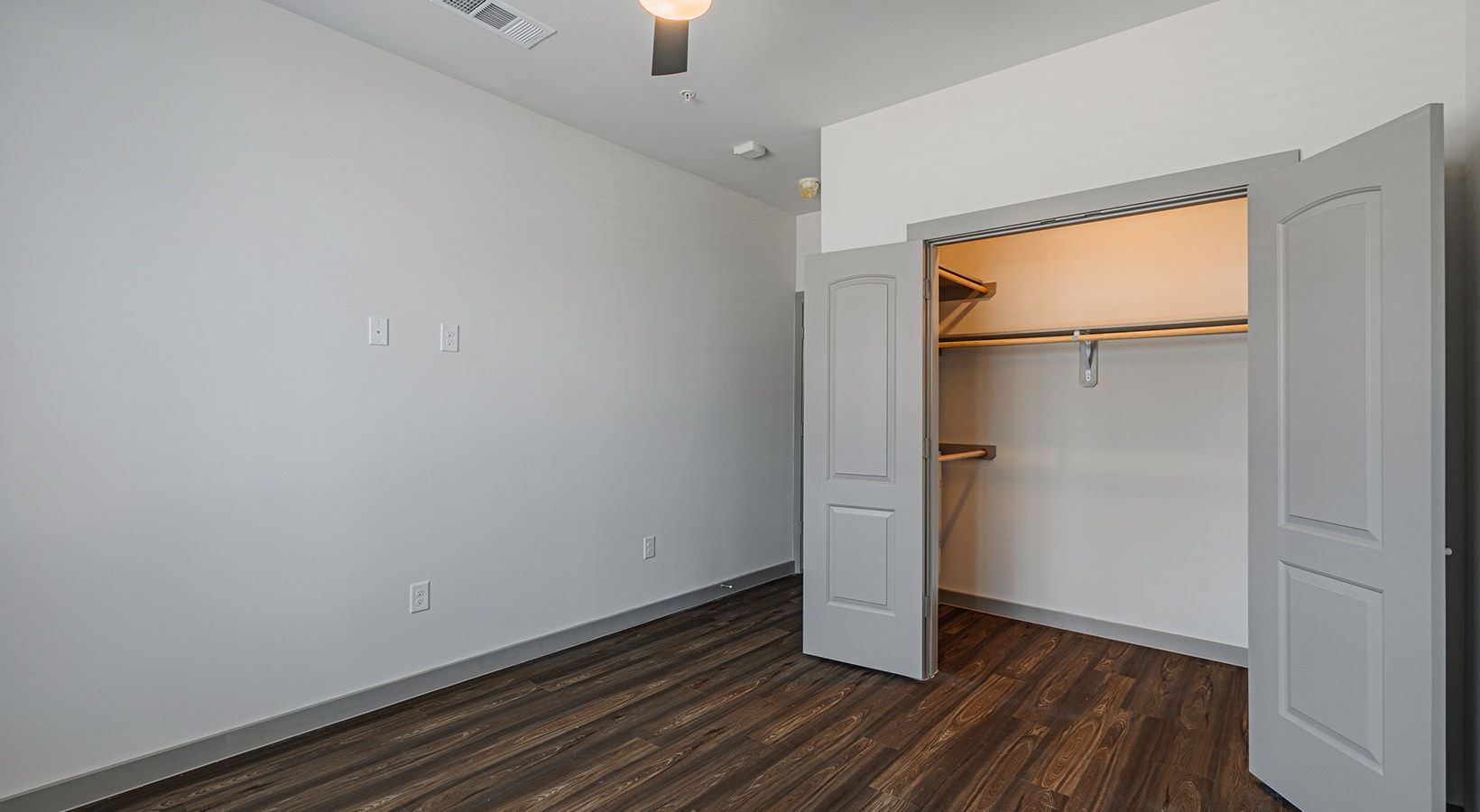 Empty bedroom with gray walls, dark wood floor, and an open closet with sliding doors.