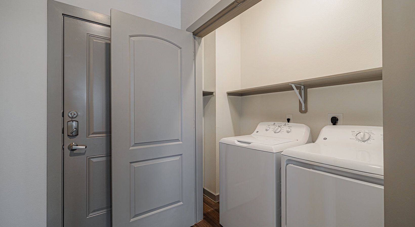 Laundry room with white washer and dryer beside a gray door and open shelving.