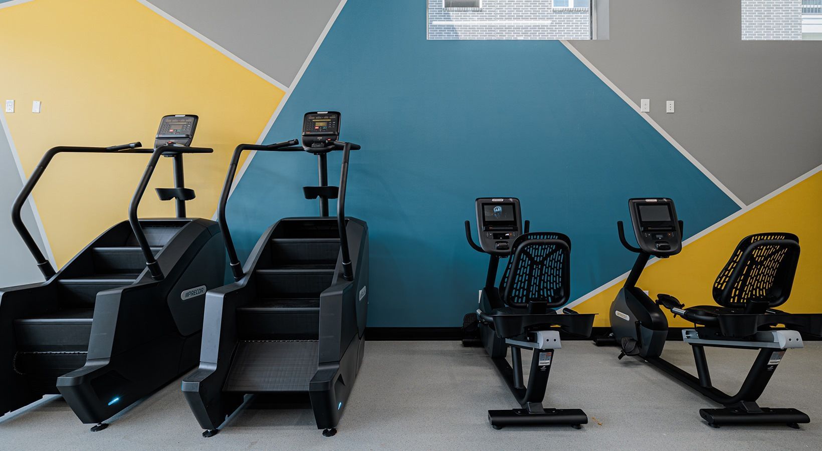 Five cardio exercise machines in a gym, mostly black with yellow and teal accent wall behind them