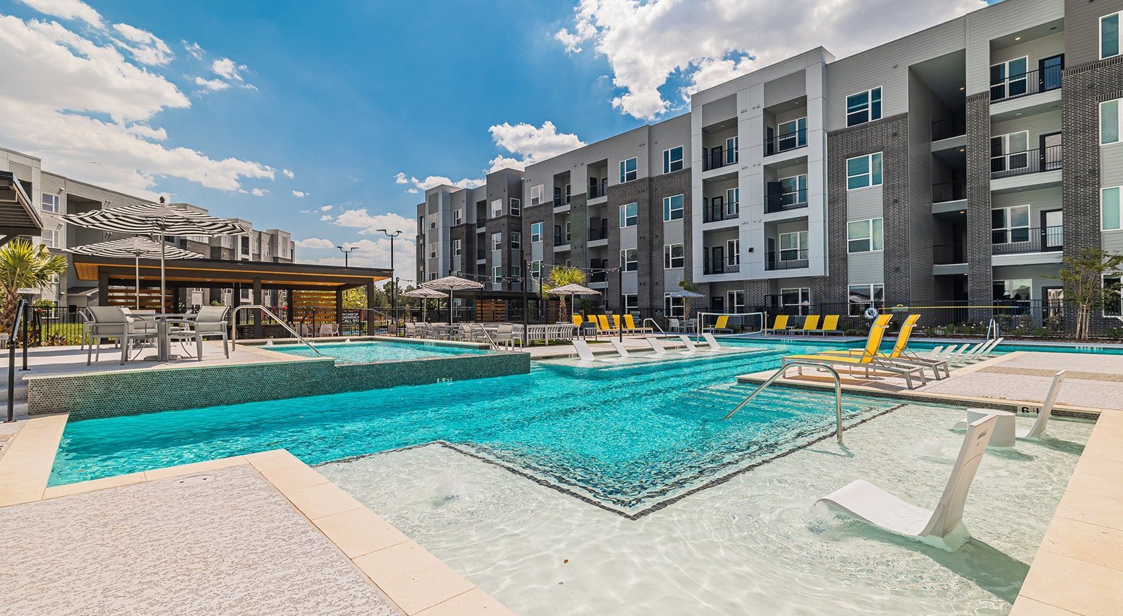 Modern apartment complex pool with turquoise water, yellow lounge chairs, and a covered seating area under a blue sky