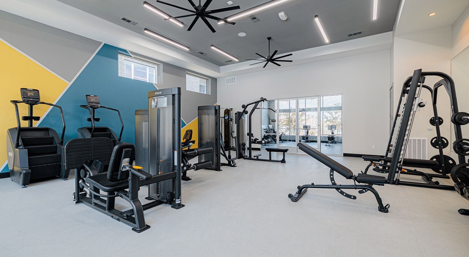 Modern gym with exercise machines, a cable bench, and large windows in a bright white room