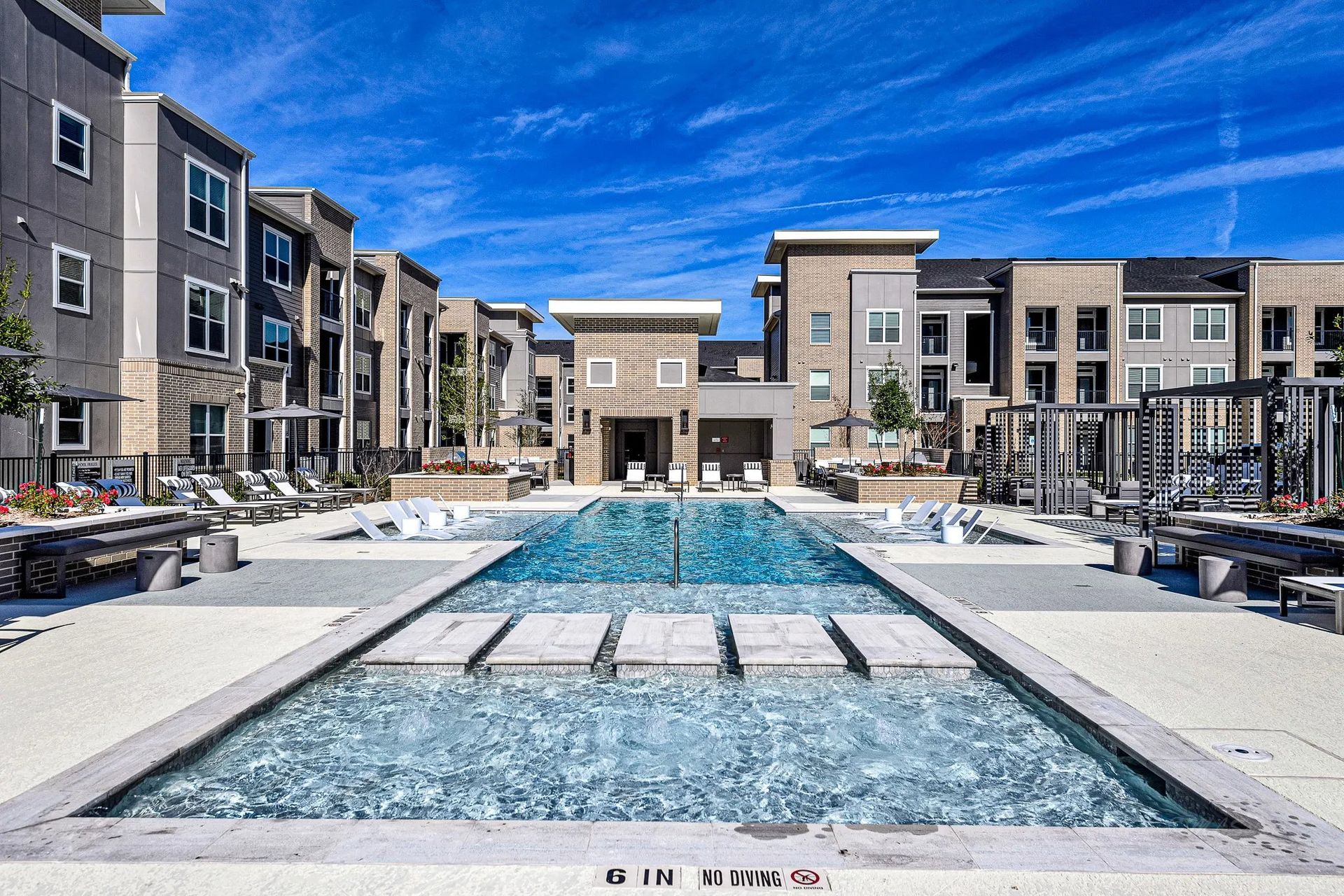 Modern apartment courtyard with a long rectangular pool and lounge chairs under a blue sky