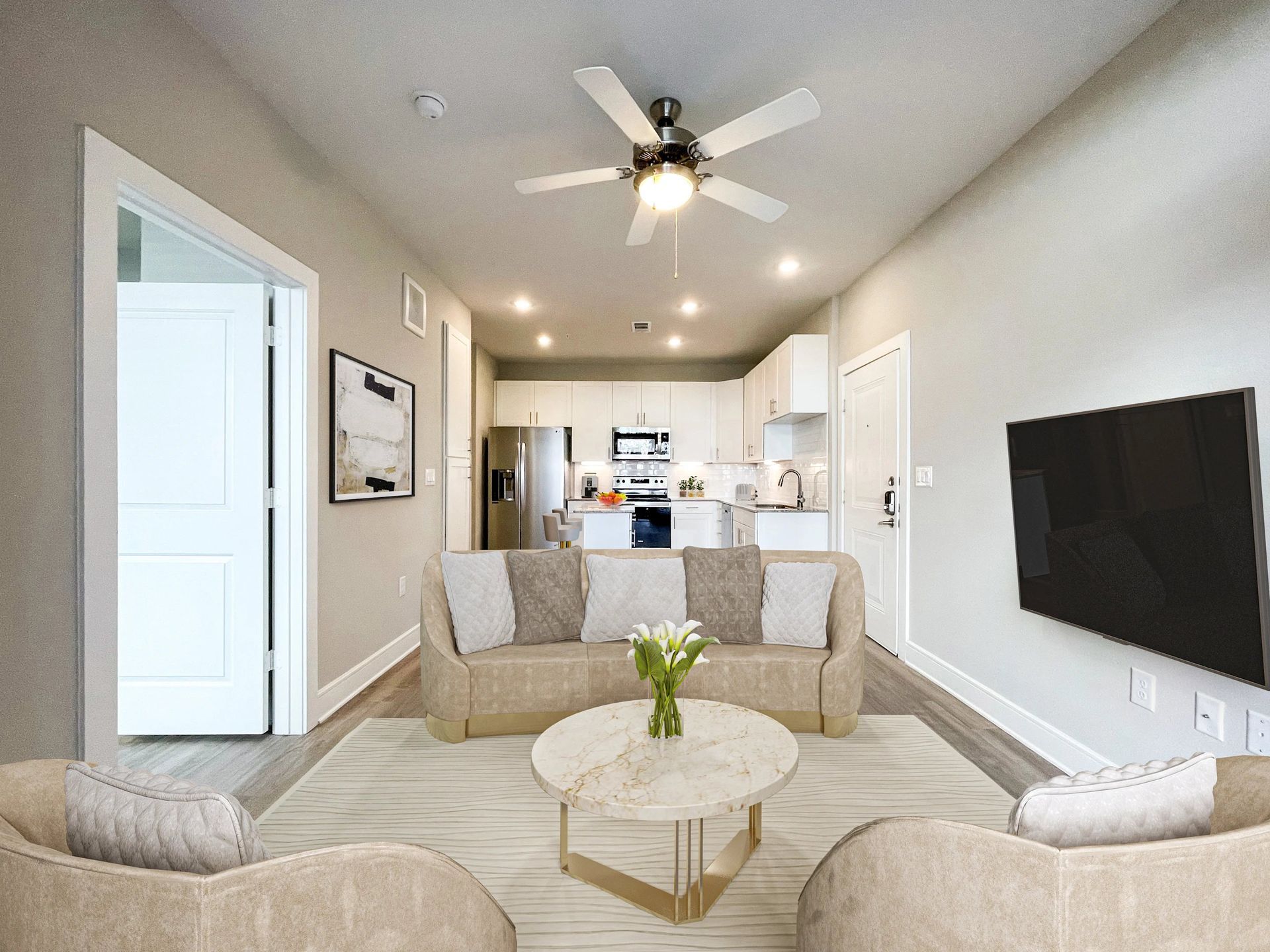 Bright modern living room with beige sofa, round coffee table, wall-mounted TV, and open kitchen in back.