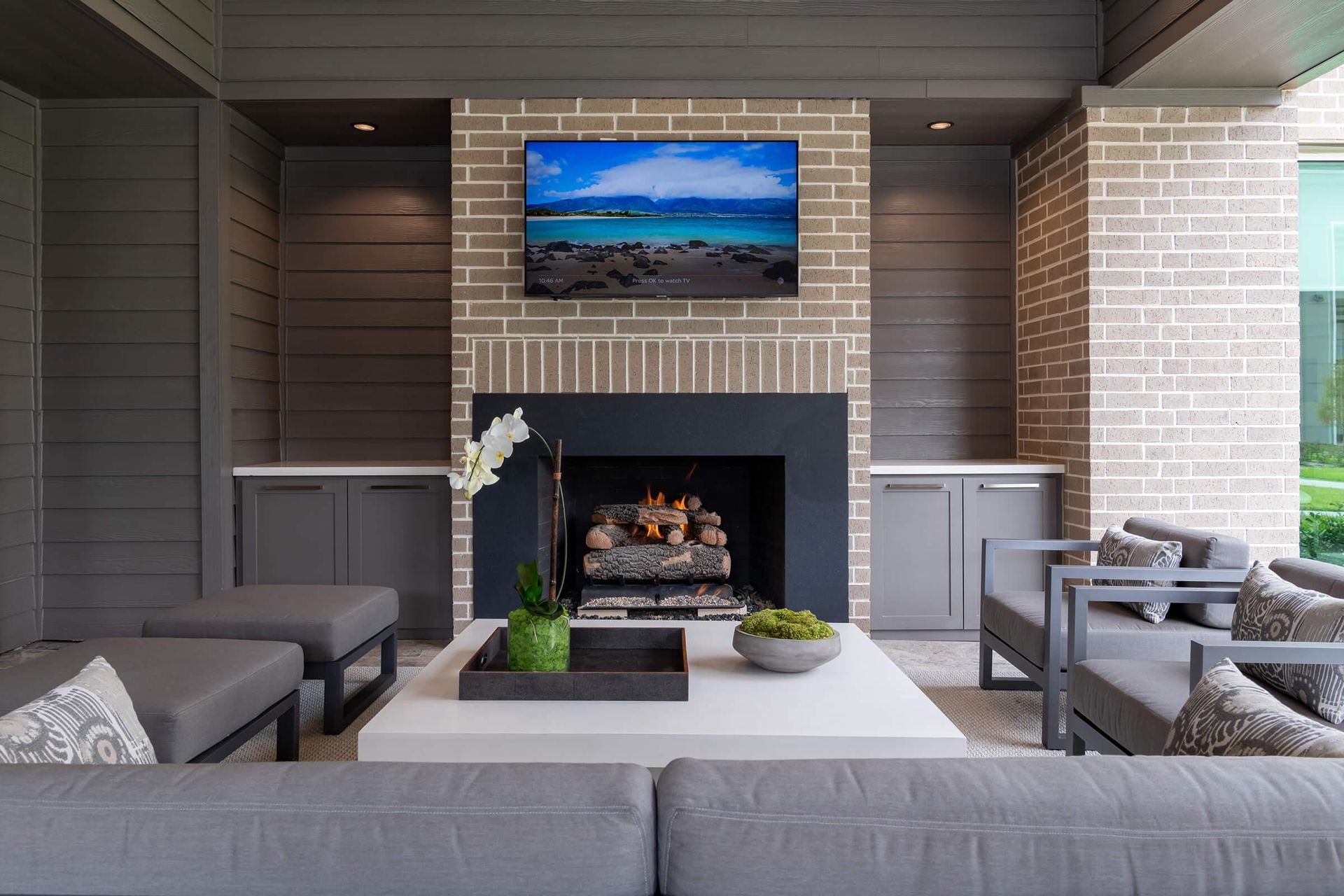 Modern outdoor patio with a brick fireplace, wall-mounted TV, and gray seating around a coffee table.