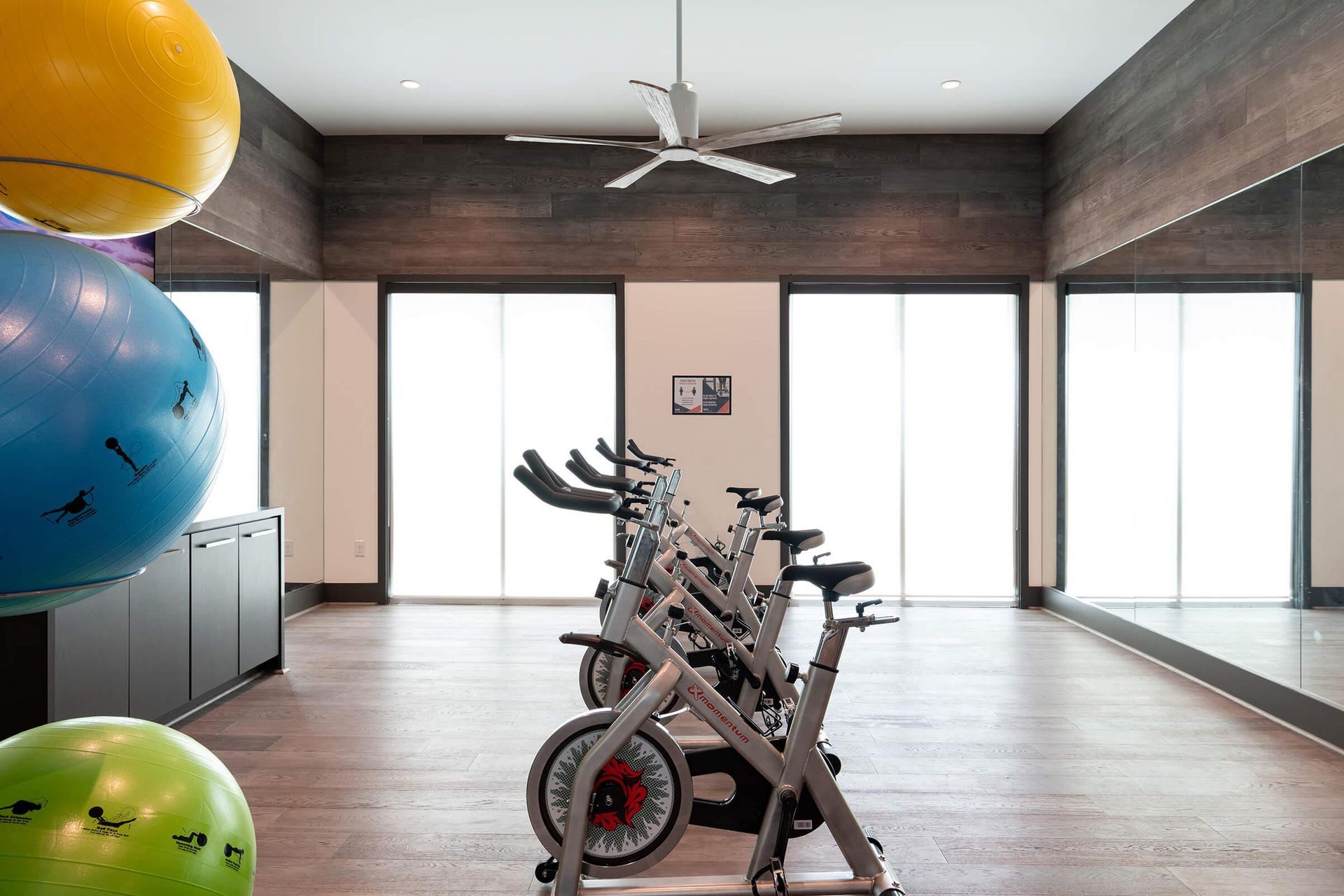 Gym with stationary bikes, floor-to-ceiling windows, mirrored wall, and colorful exercise balls on the left.
