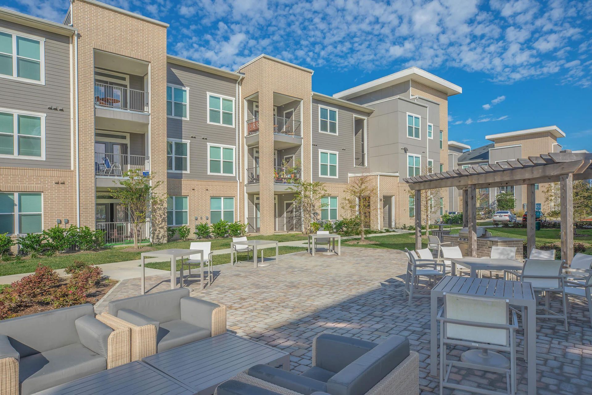 Apartment courtyard with lounge chairs, pergola dining area, and a three-story building under blue sky