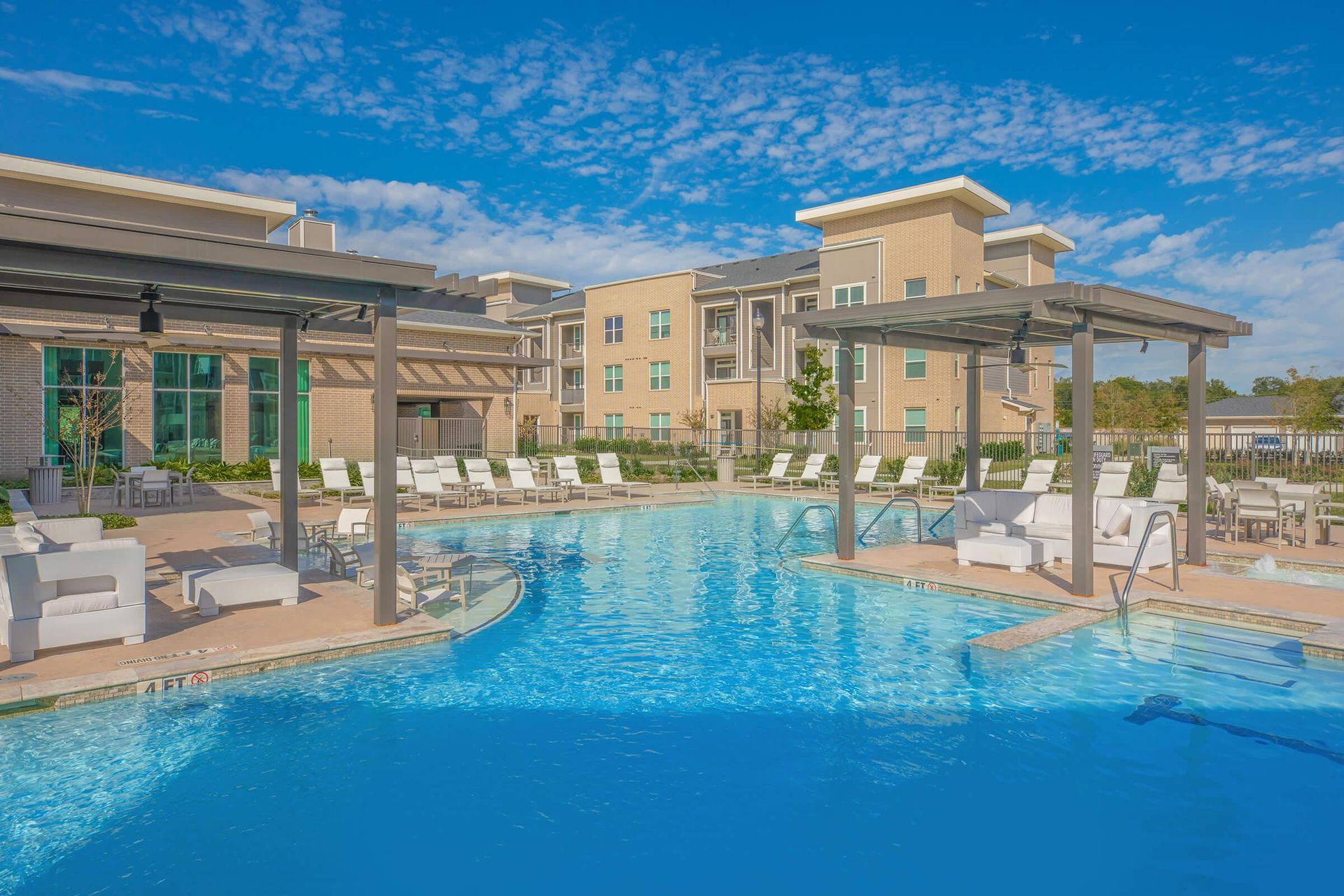 Resort-style outdoor pool with lounge chairs, cabanas, and a hotel building under a blue sky