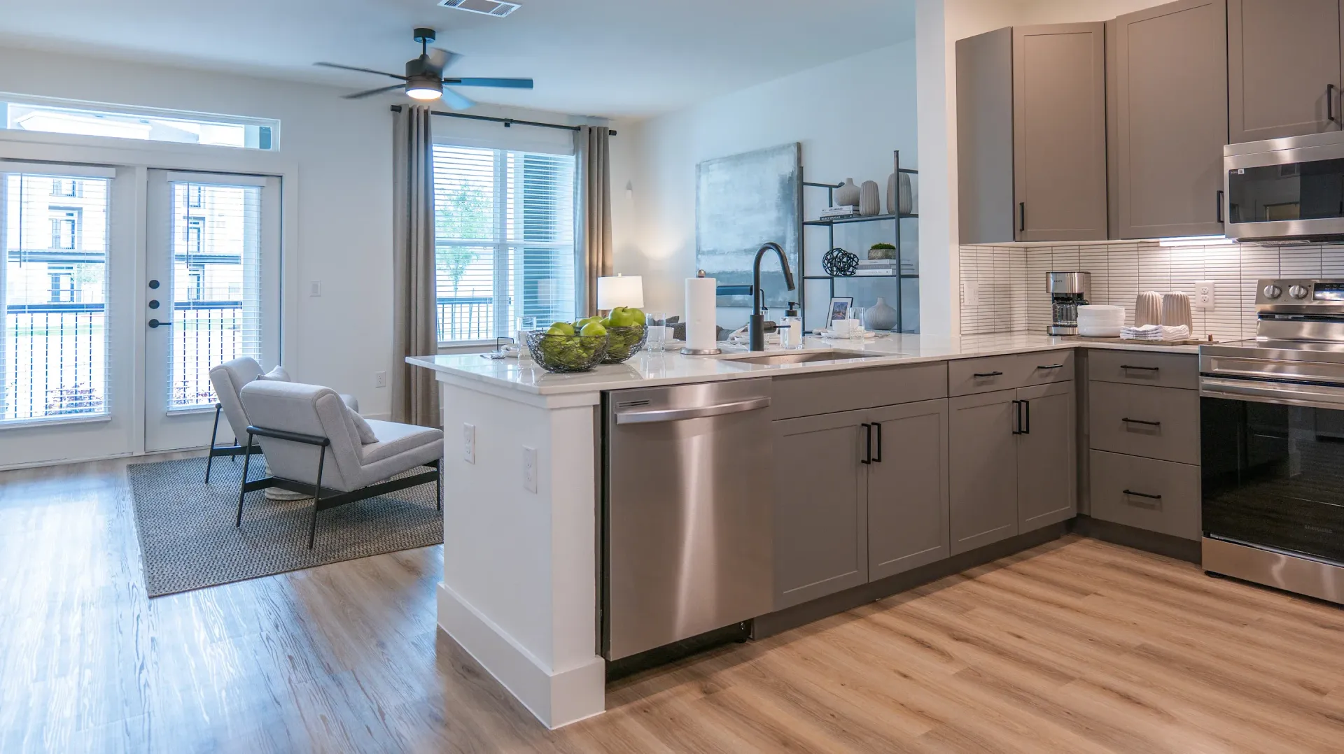Bright modern open-plan kitchen and living room with gray cabinets, island, hardwood floors, and white chairs by windows