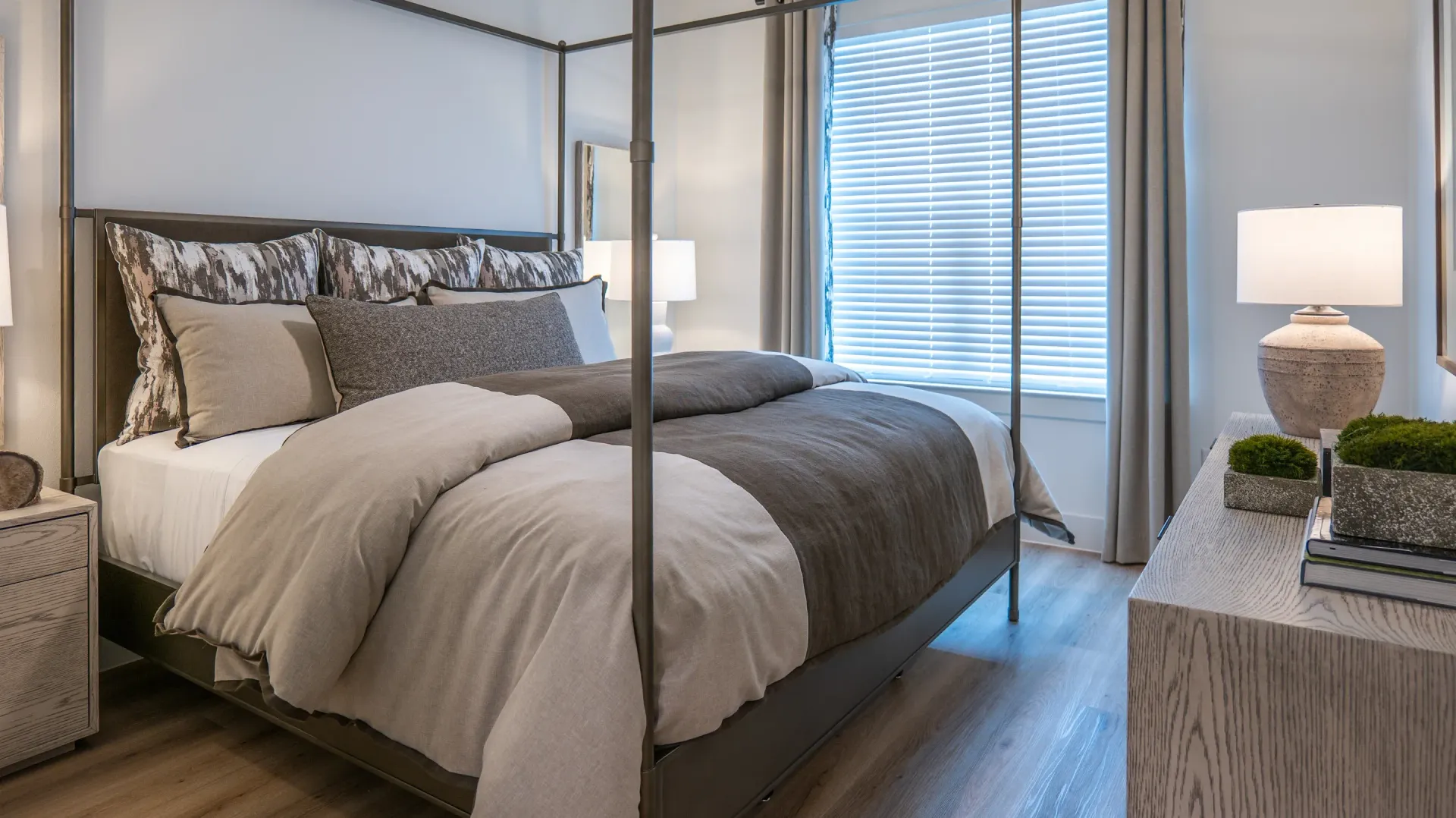 Modern bedroom with a four-poster bed, gray bedding, and a bedside lamp near a window with blinds