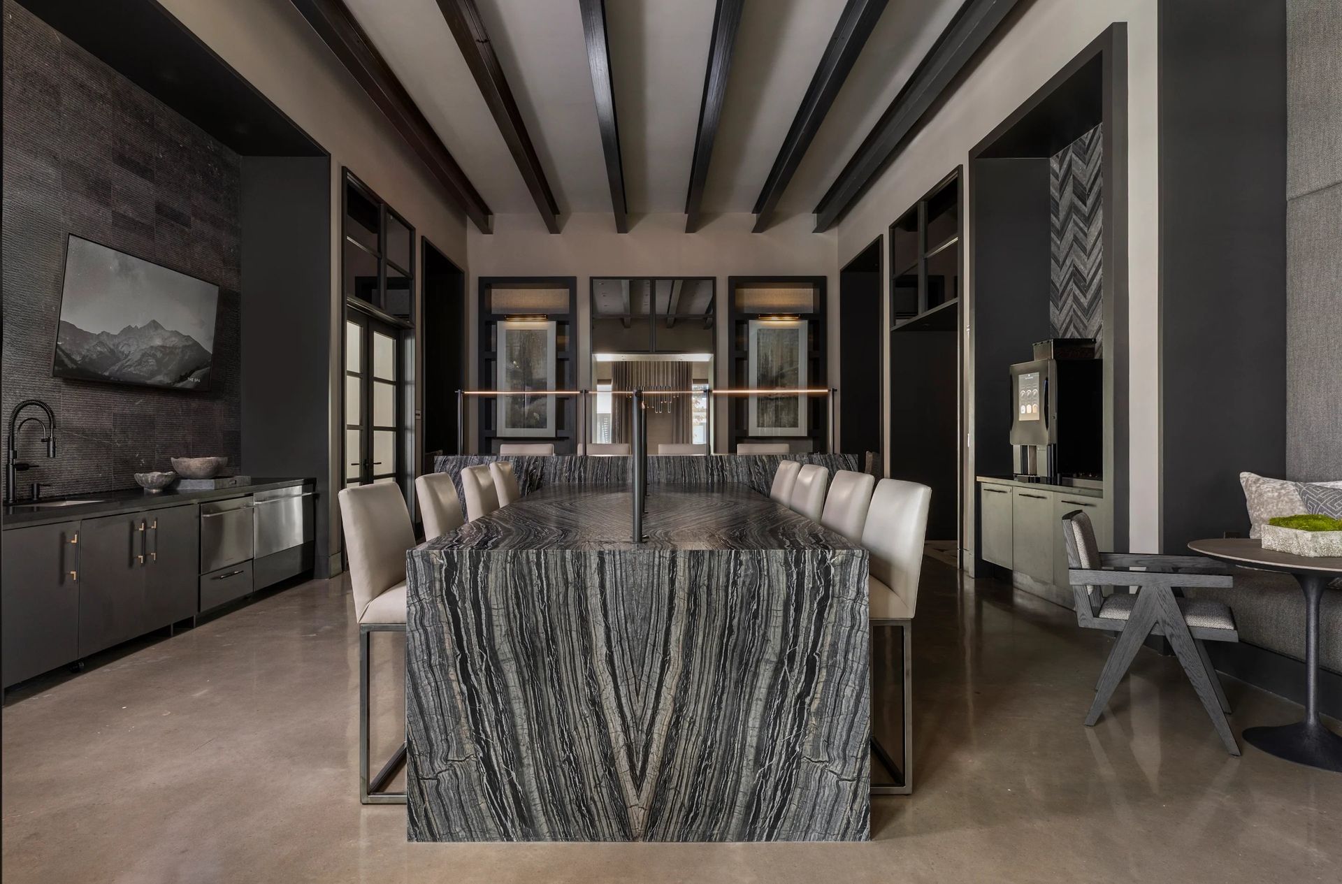 Modern dining room with a long table, upholstered chairs, exposed ceiling beams, and dark decor.