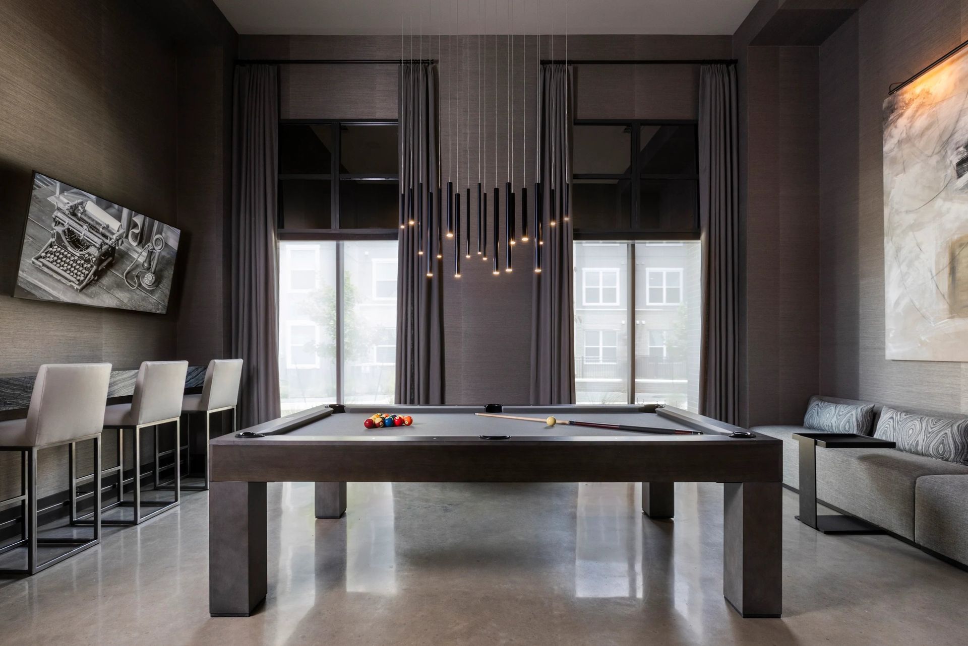 Modern game room with a pool table, gray walls, bar stools, and large windows