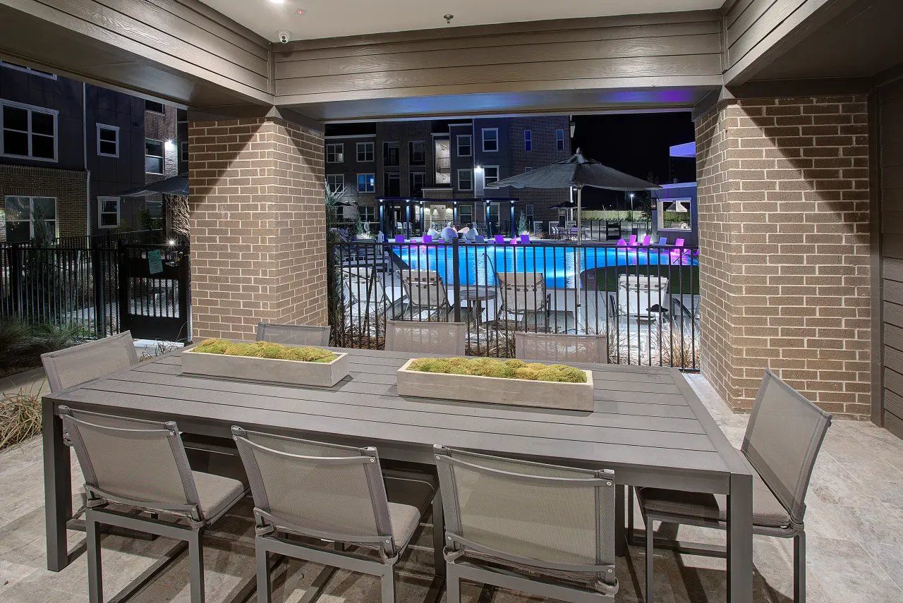 Covered patio dining area overlooking a lit pool at night with a table and chairs