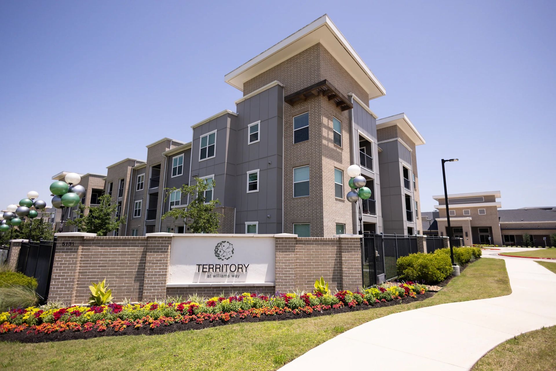 Modern gray apartment building with flower beds and a sidewalk under a clear blue sky