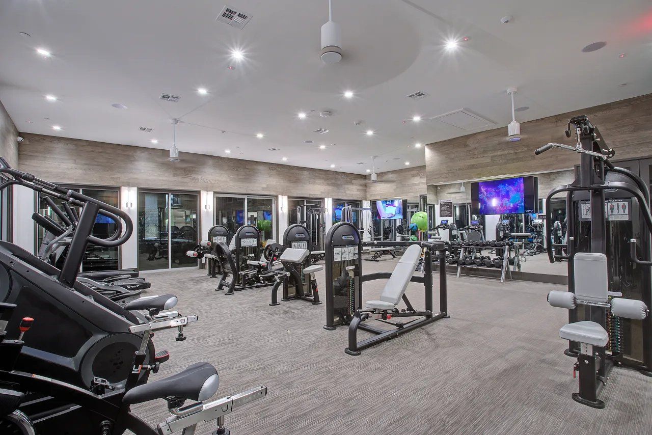 Modern gym interior with cardio and weight machines, mirrors, bright ceiling lights, and a TV screen