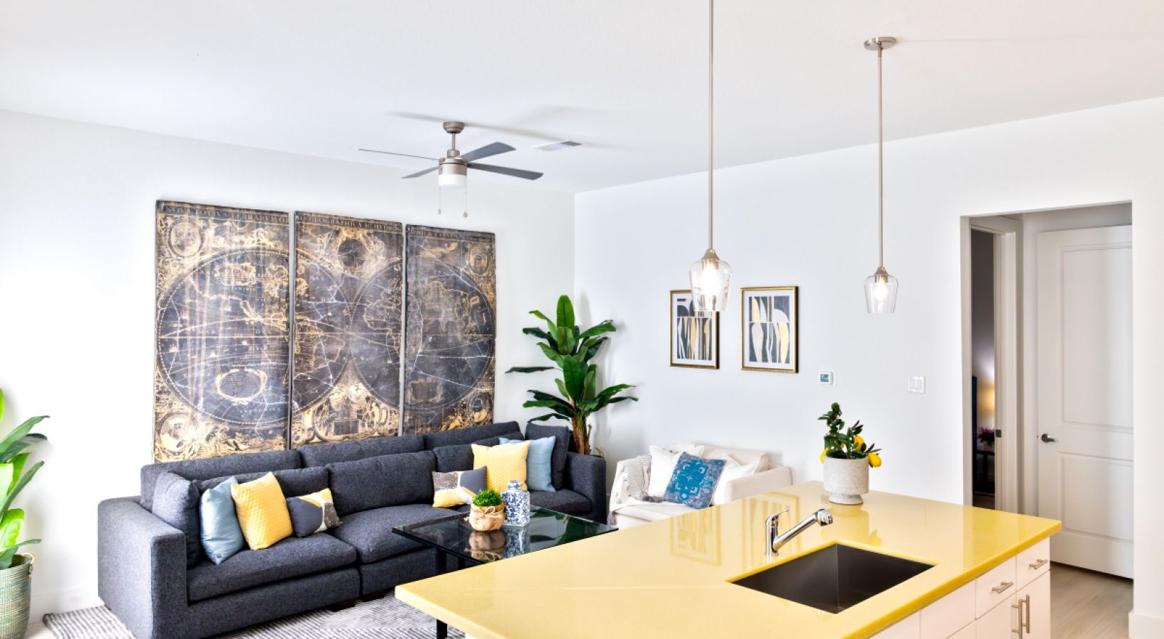 Bright modern living room with dark sofa, yellow accents, large artwork, and a wooden dining table