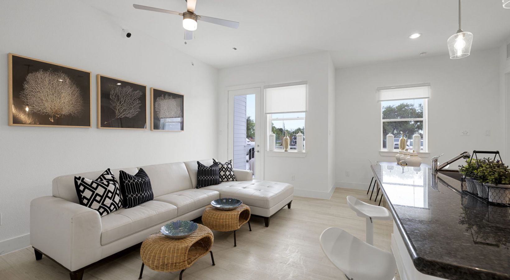 Bright modern living room with white sofa, patterned pillows, ottoman, wall art, and kitchen counter nearby