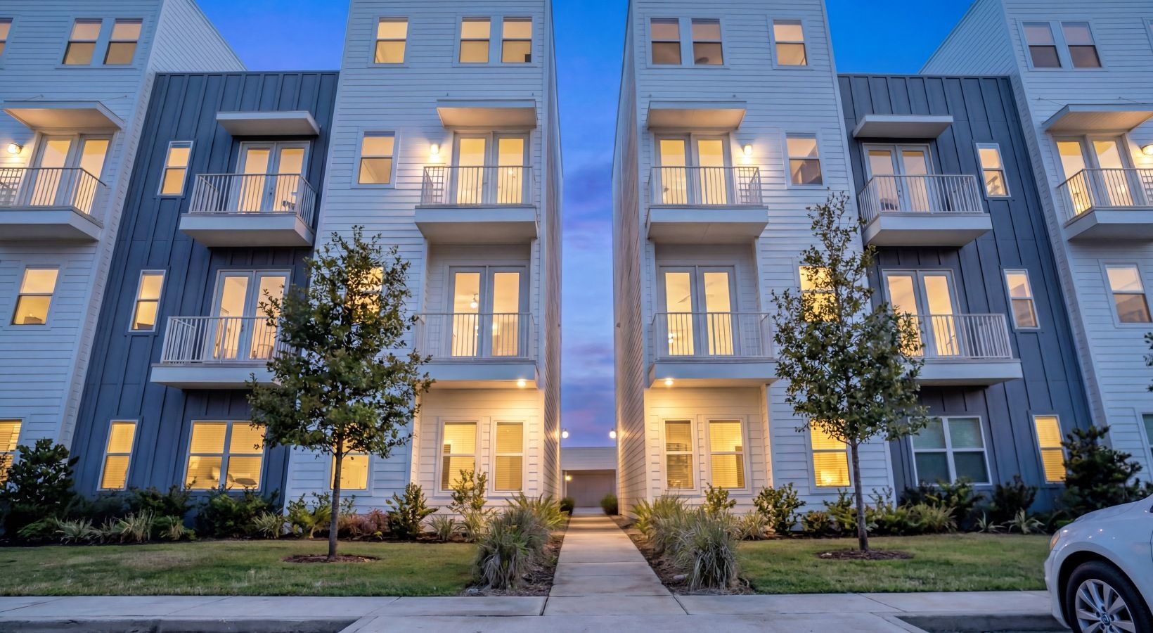 Modern apartment building exterior at dusk with lit windows and a sidewalk entrance