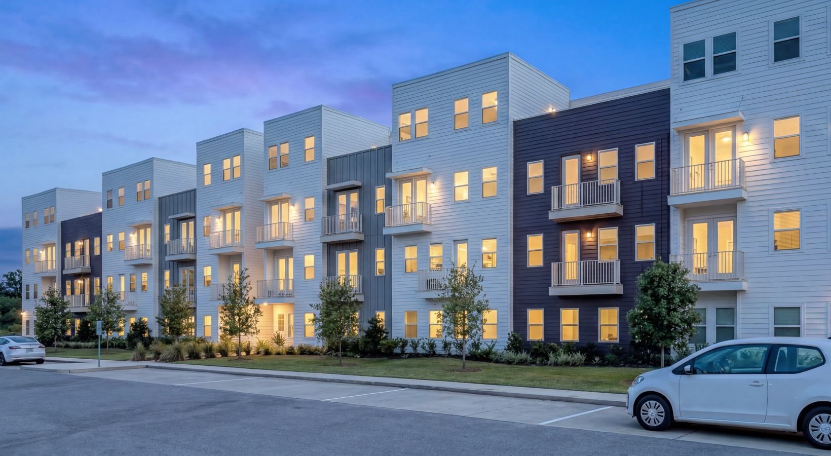 Modern apartment building exterior at dusk with lit windows and parked cars in front