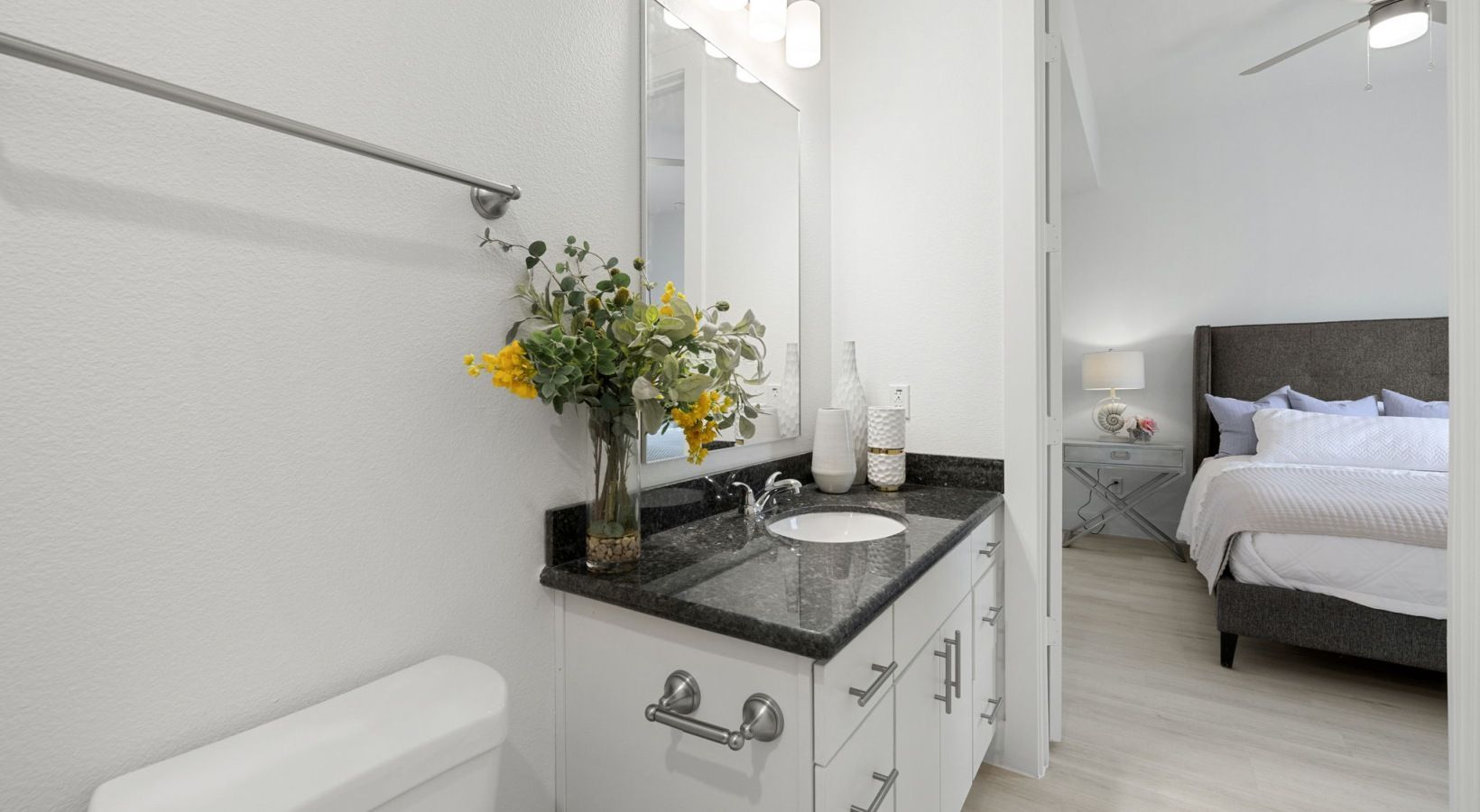 Bright bathroom vanity with flowers, mirror, and a glimpse of a bedroom beyond the doorway