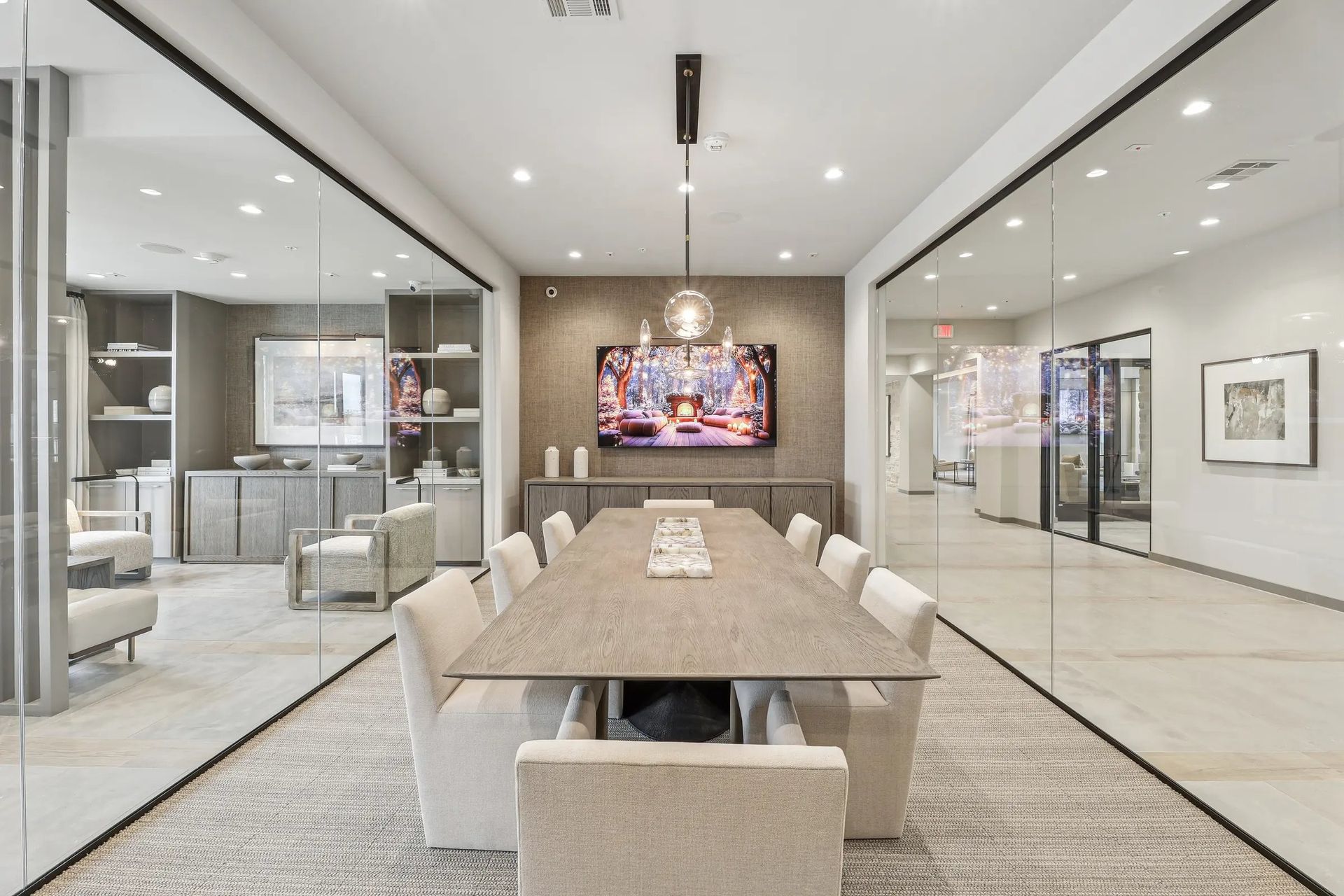 Modern dining room with a long table, beige chairs, glass walls, and a wall-mounted TV in the background