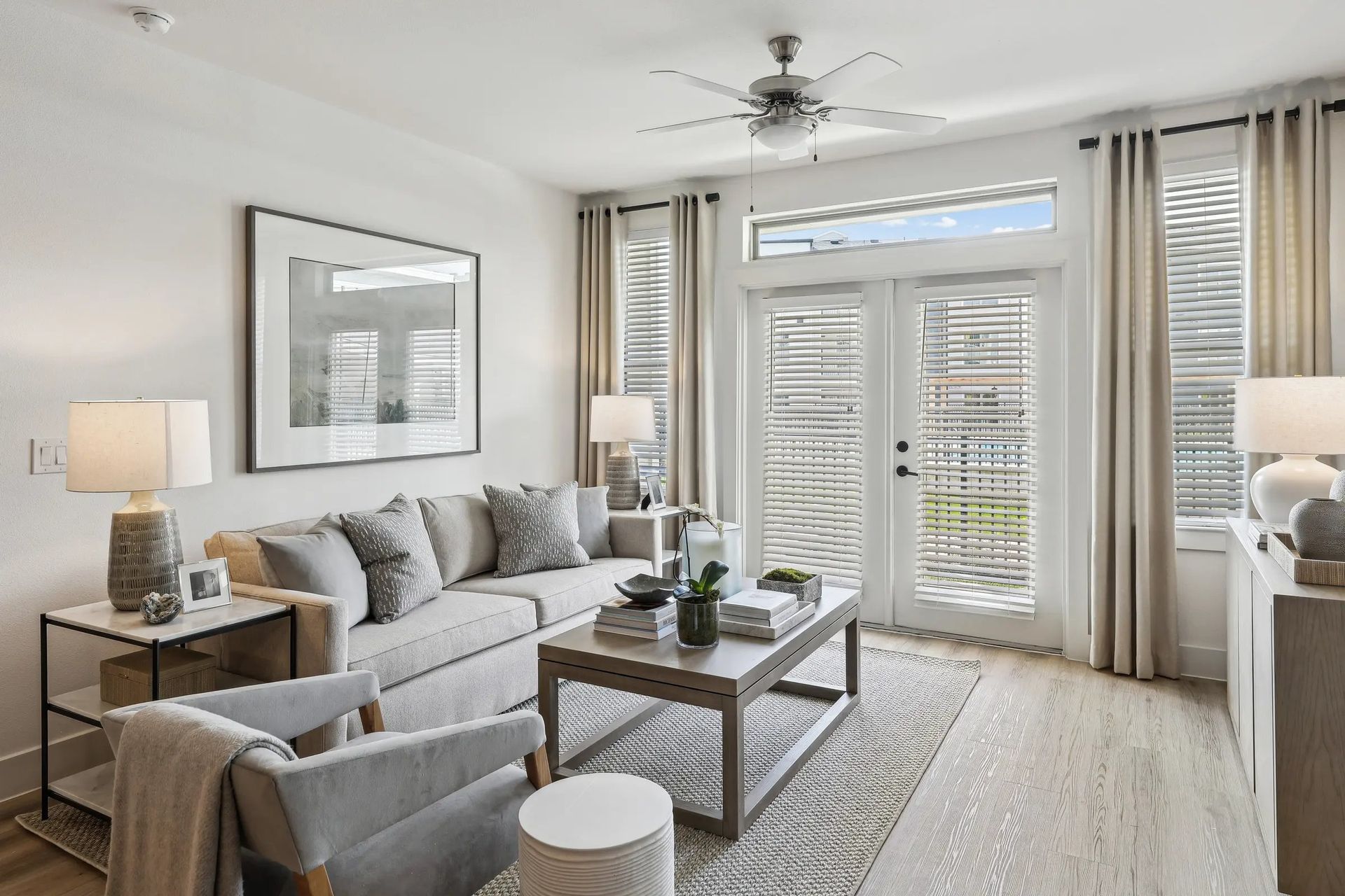 Bright living room with gray sofa, glass coffee table, lamp, and French doors with shutters and curtains
